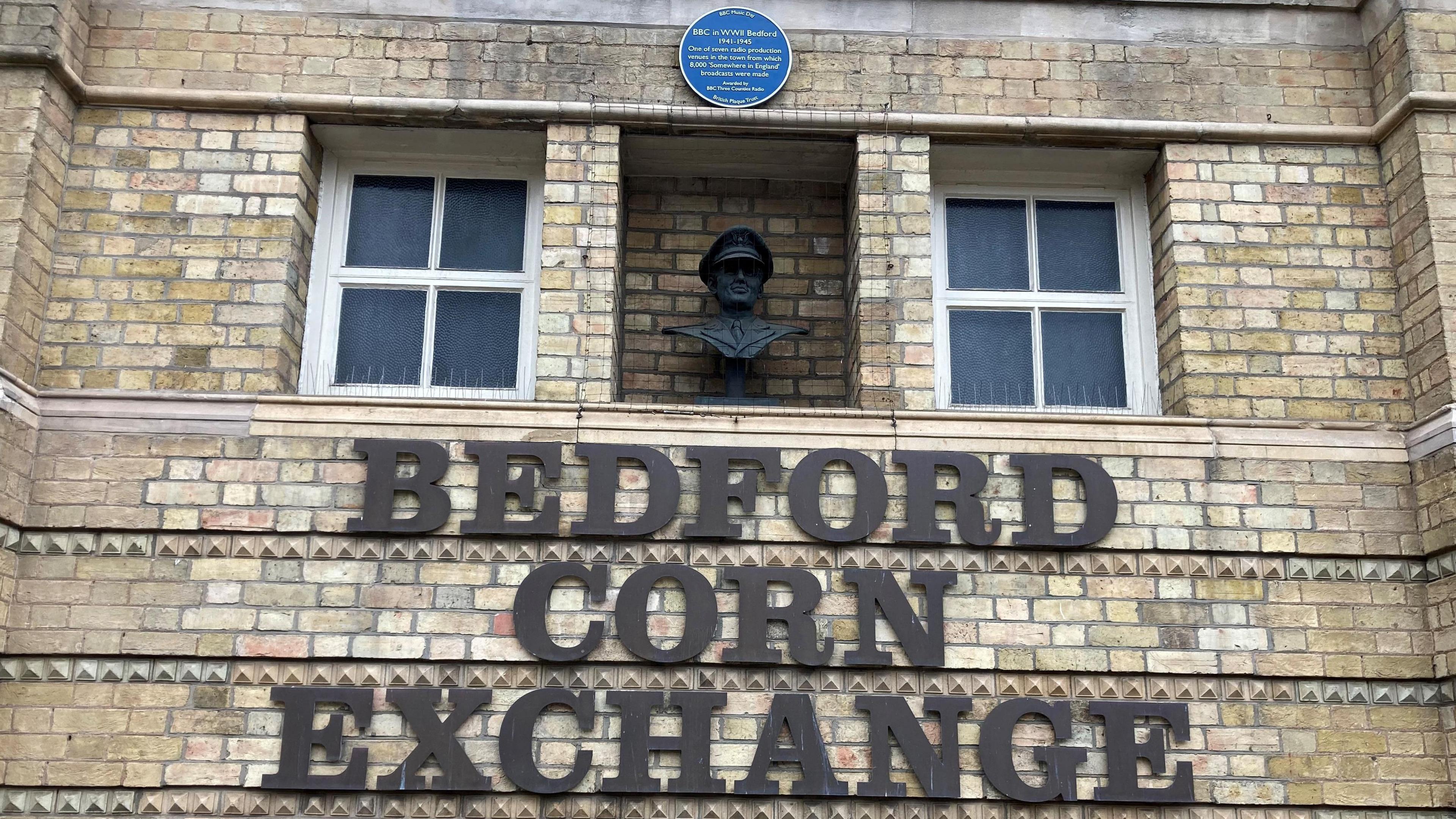 A blue plaque and statue on the front of Bedford Corn Exchange. The statue is of his head, set back into the wall, which is made of light brick. There are two white windows and signage for the building. 