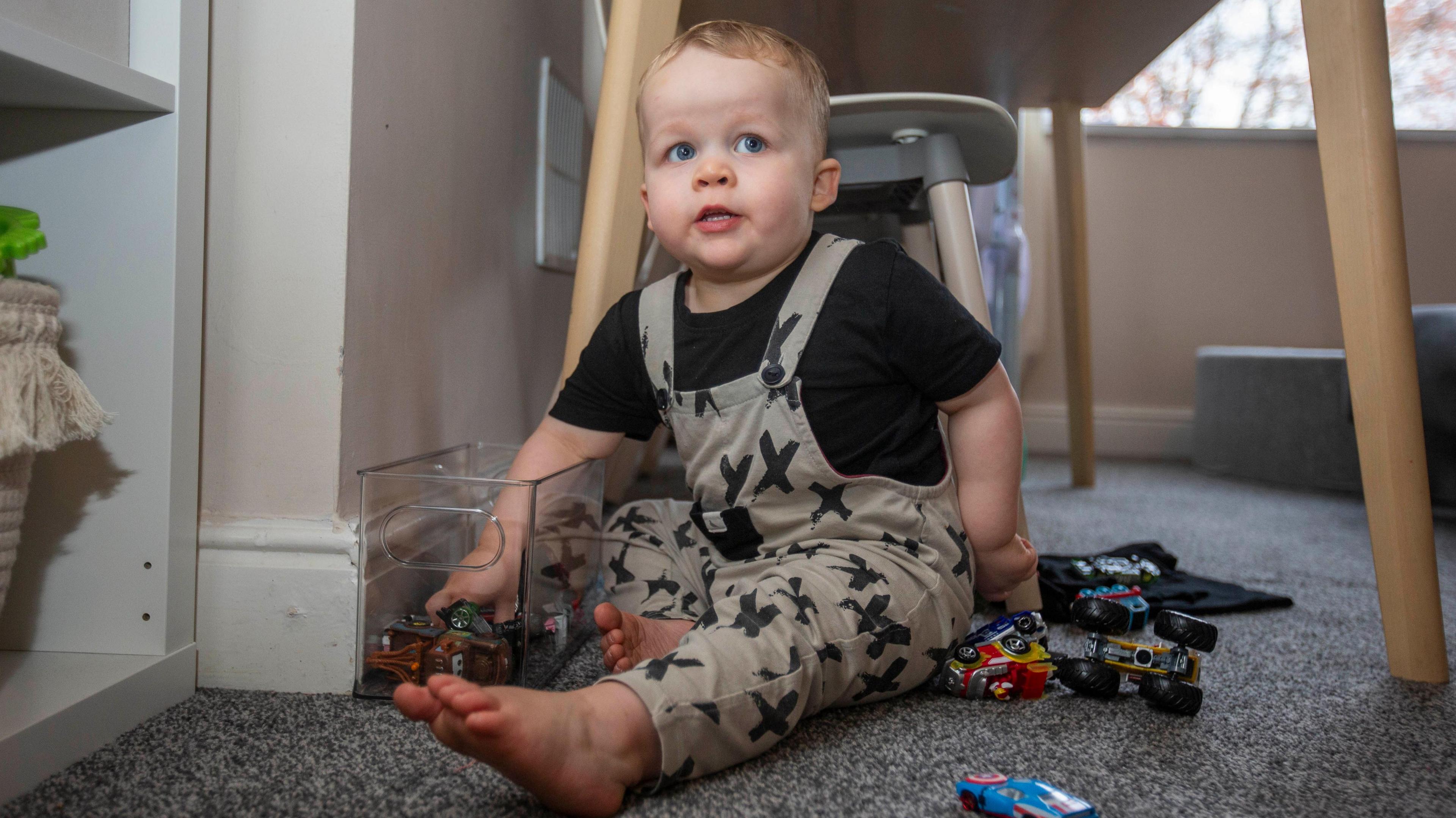 A little boy sat on grey carpet under a table. He is surrounded by toy cars and has his hand in a plastic tub of toy cars, with a cheeky expression on his face.