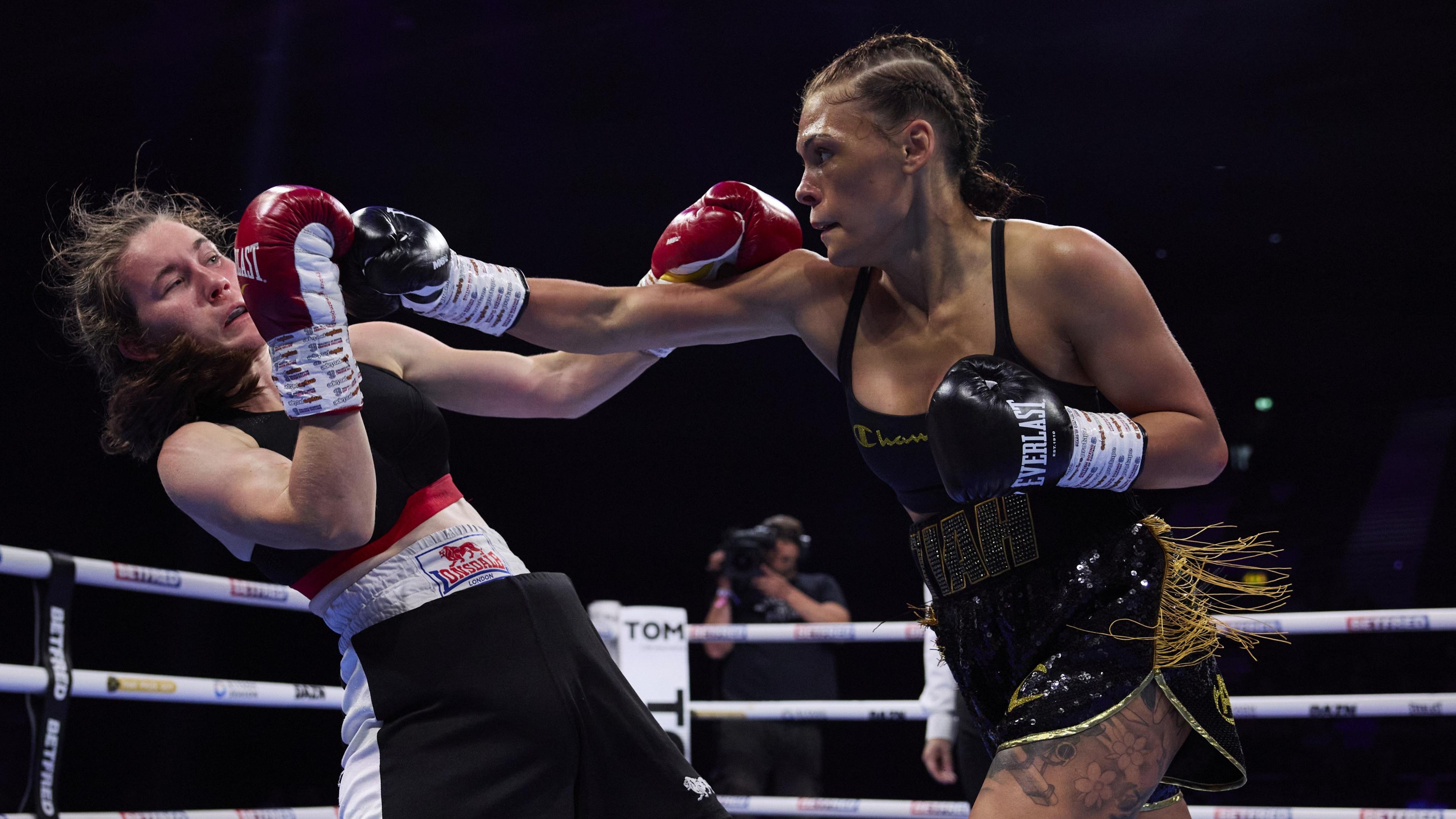 Tiah throwing a punch towards her opponent in the boxing ring. She is wearing black shorts with gold fringing details and black boxing gloves, with her hair in tight plaits.