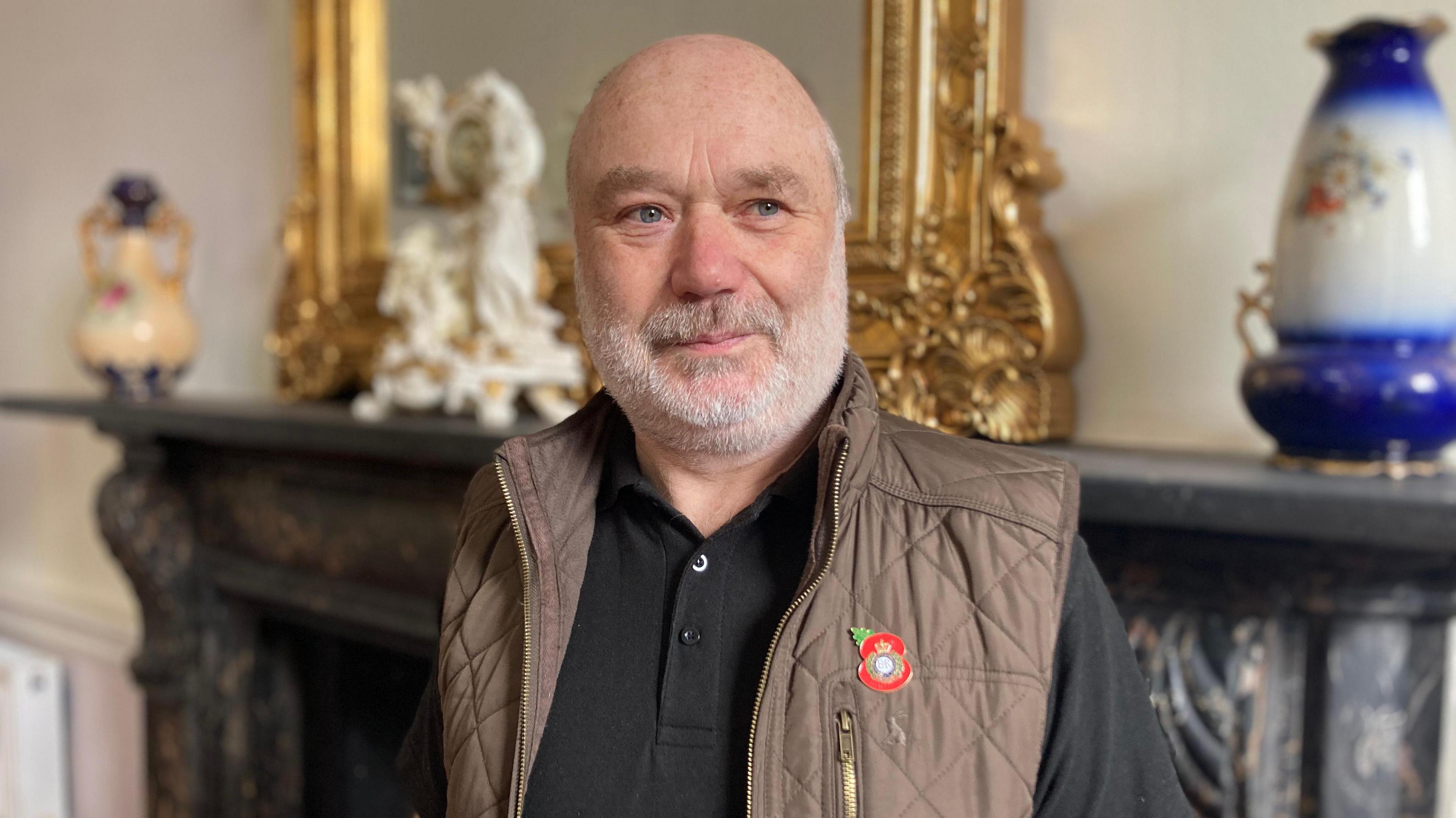 John Naylor has a grey short beard and is smiling at the camera.  He wears a black polo shirt and brown gillet with a metal poppy on the breast pocket.  He stands in front of an ornate fireplace.