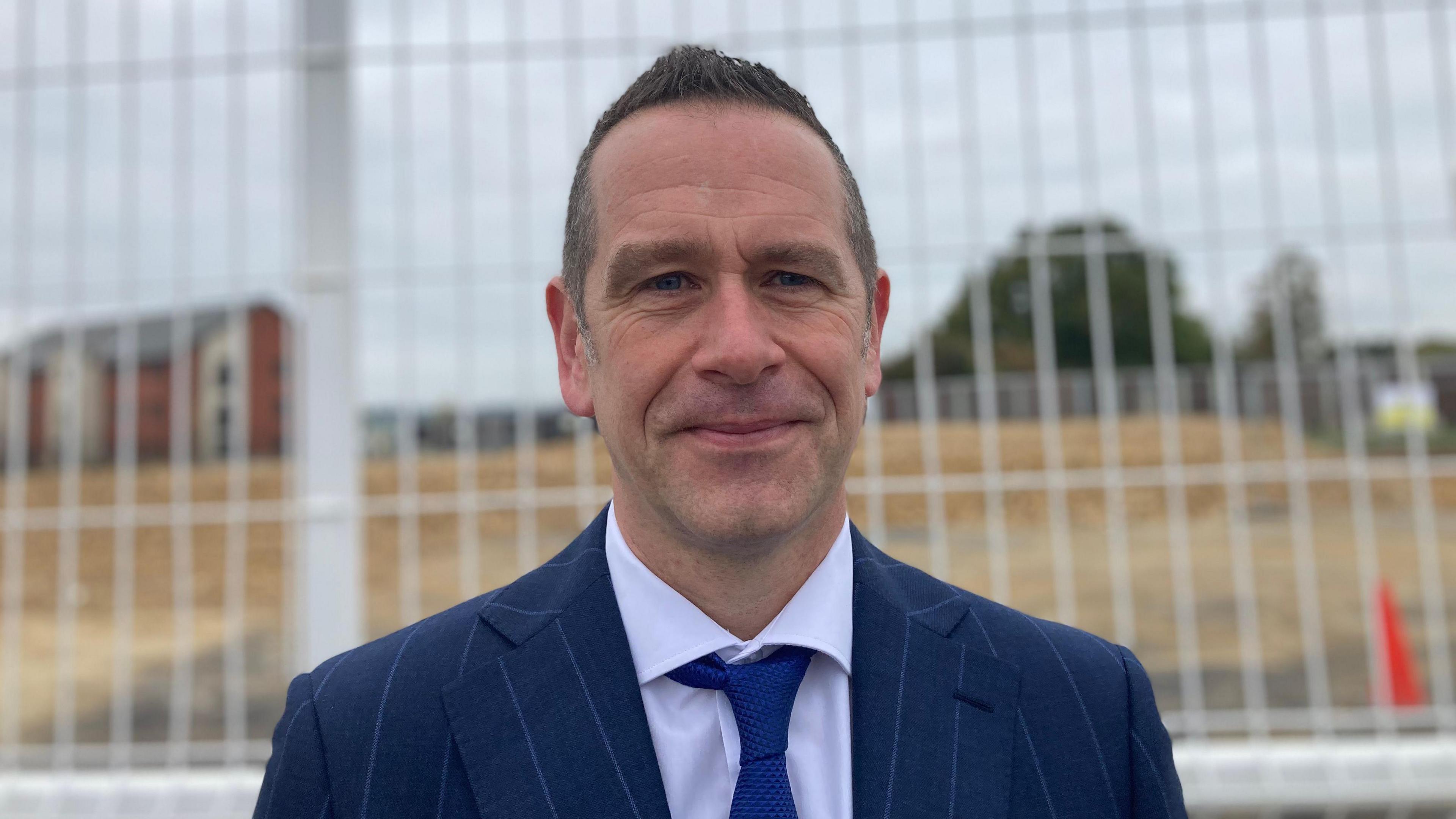 A man in a suit looking at the camera with a soft smile. He is wearing a navy striped suit and standing in front of a building site with a gate behind him.
