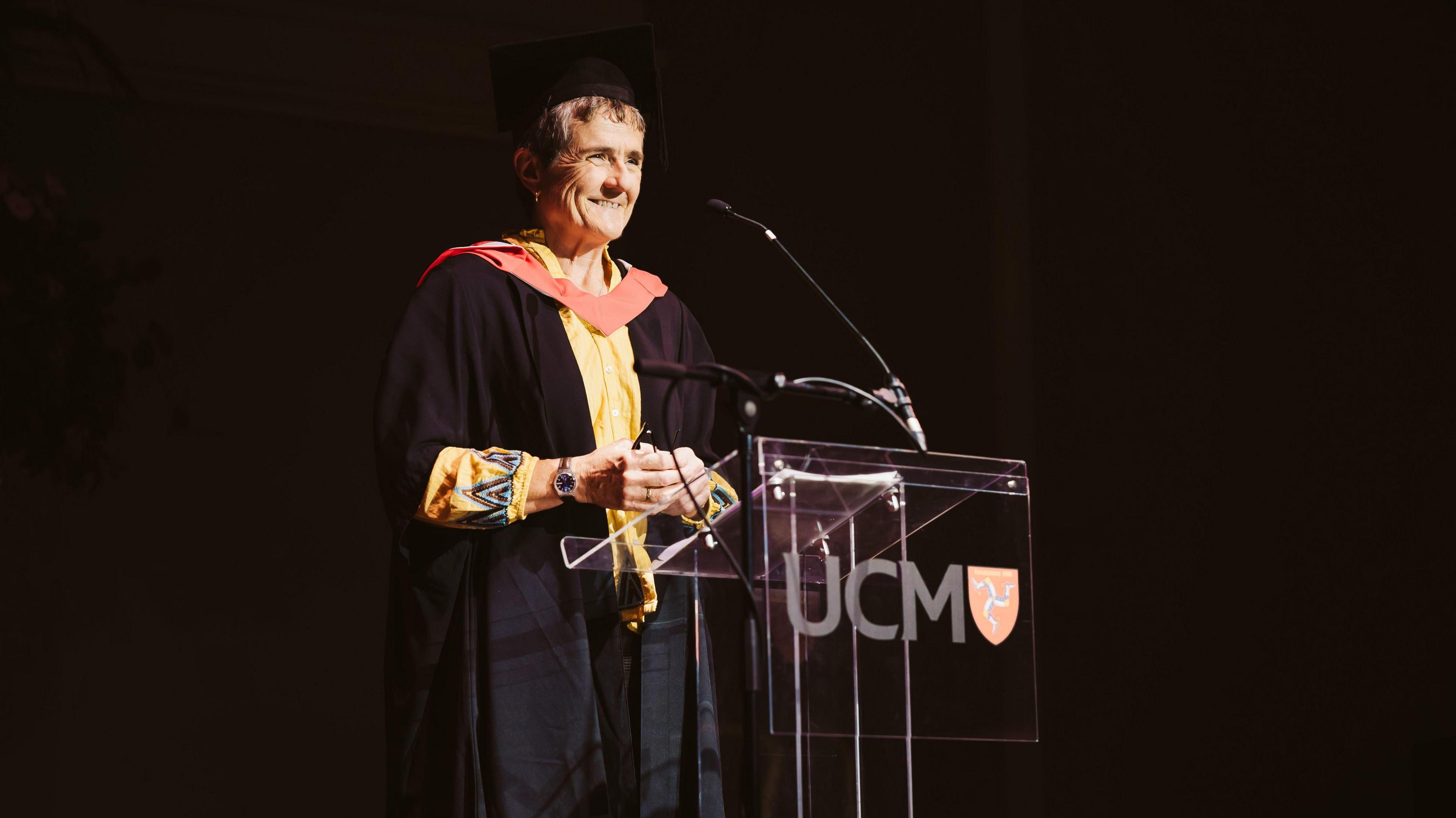 Gail Corrin a woman with short hair, wears a graduation robe and hat as she makes a speech in front of a Perspex podium with UCM on the front.  