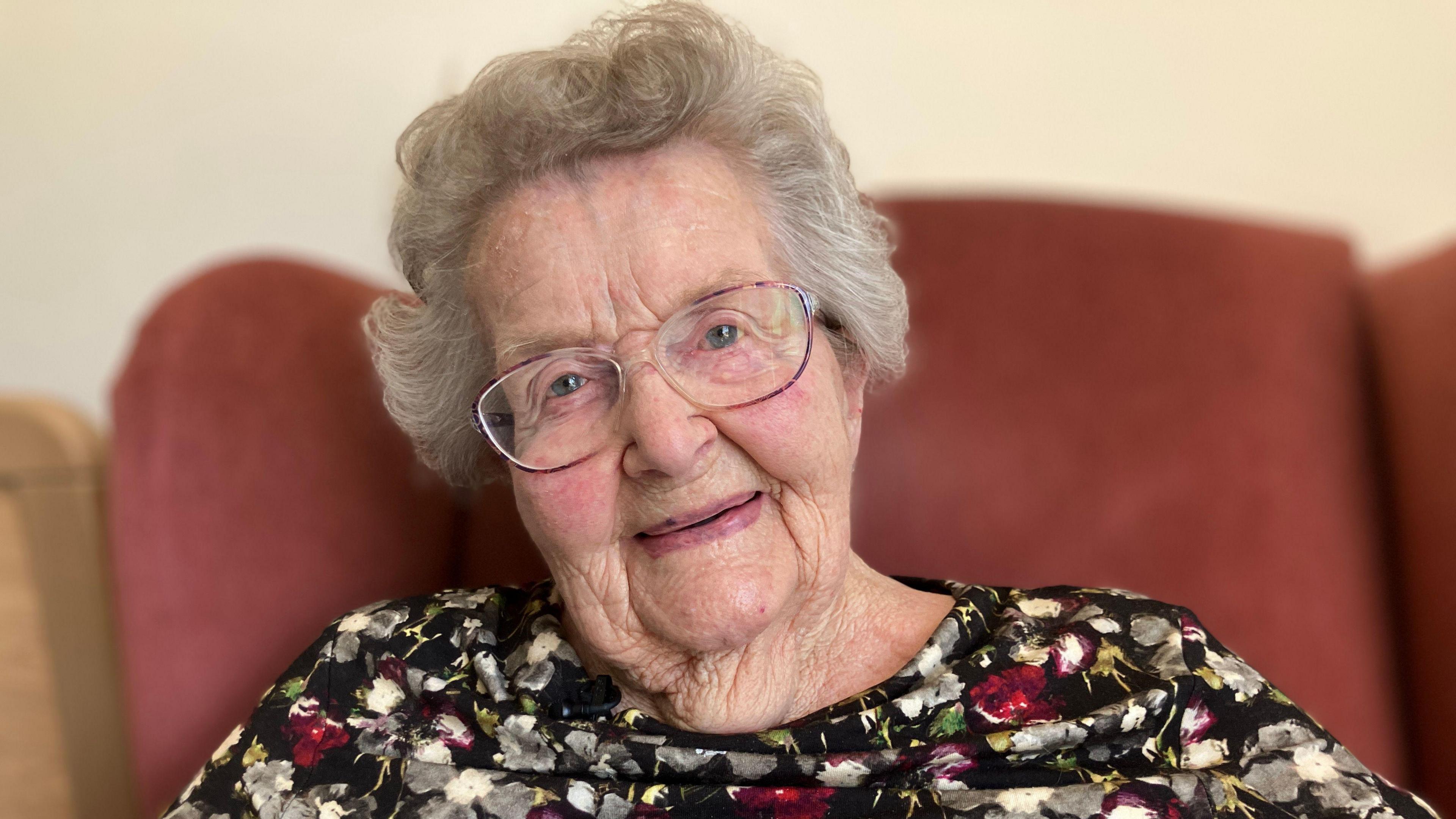 Ula Rigg sitting in a red armchair. She has round glasses on and grey hair, and a floral top