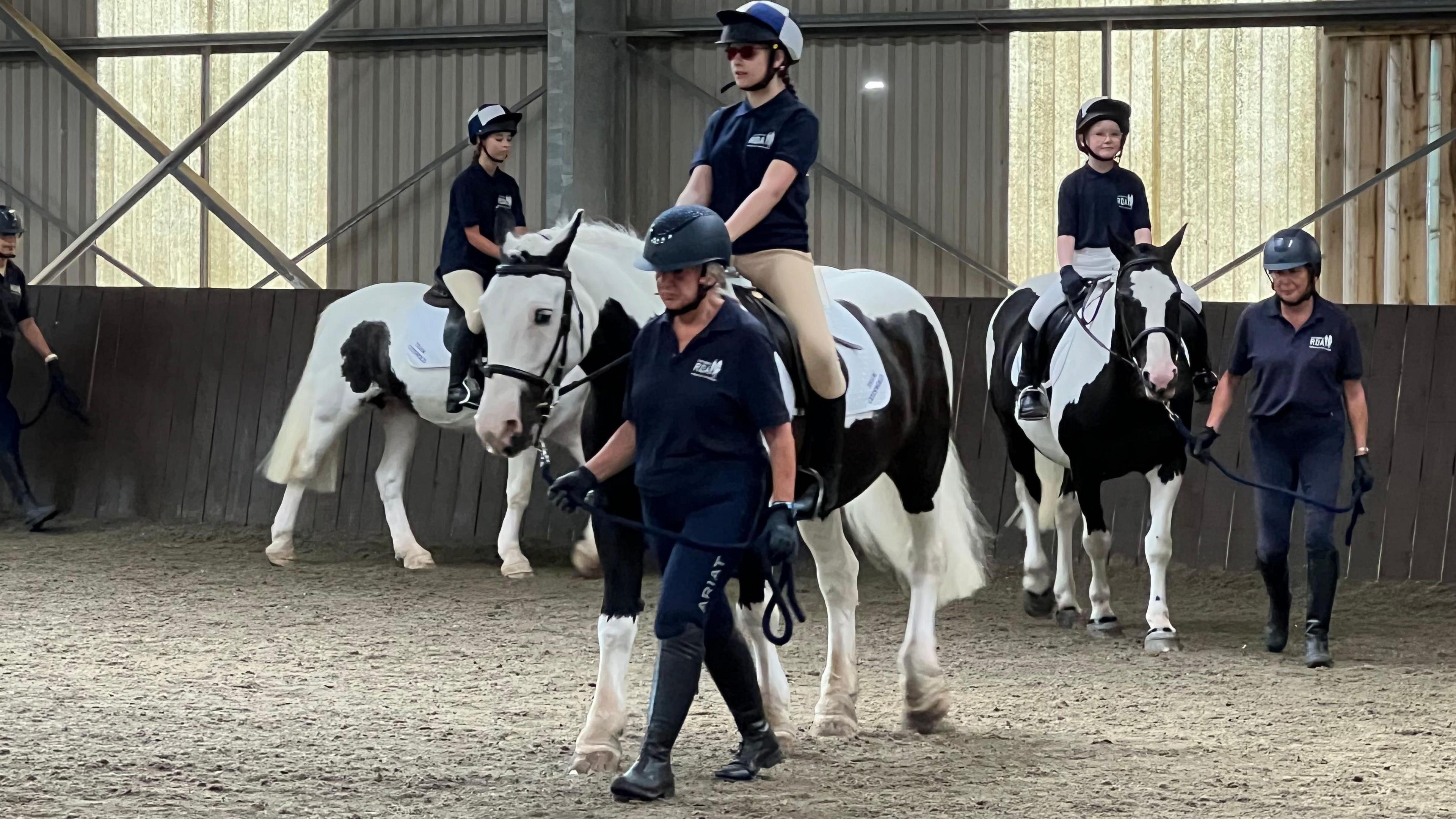 A group of three young people are led on black and white piebald ponies around an indoor riding arena by instructors.