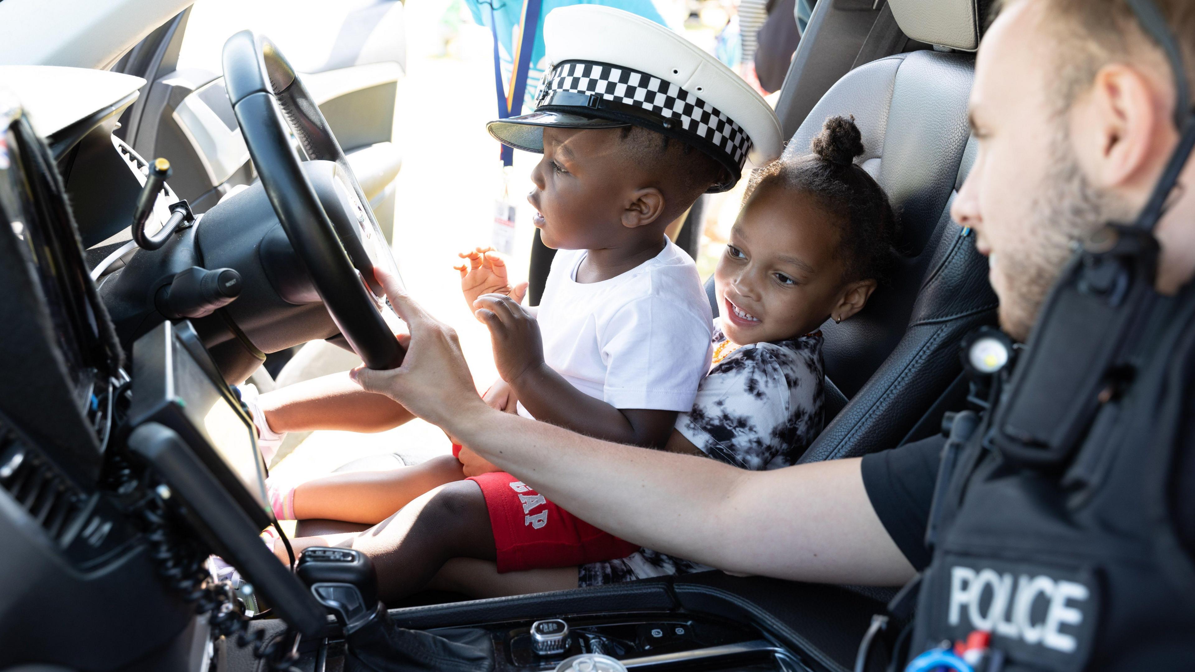 A police officer wearing their uniform, leaning over from the passenger's seat in a police car and touching the steering wheel. In the driver's seat there are two small children. The boy is wearing a white police hat and is sitting on a young girl's lap.