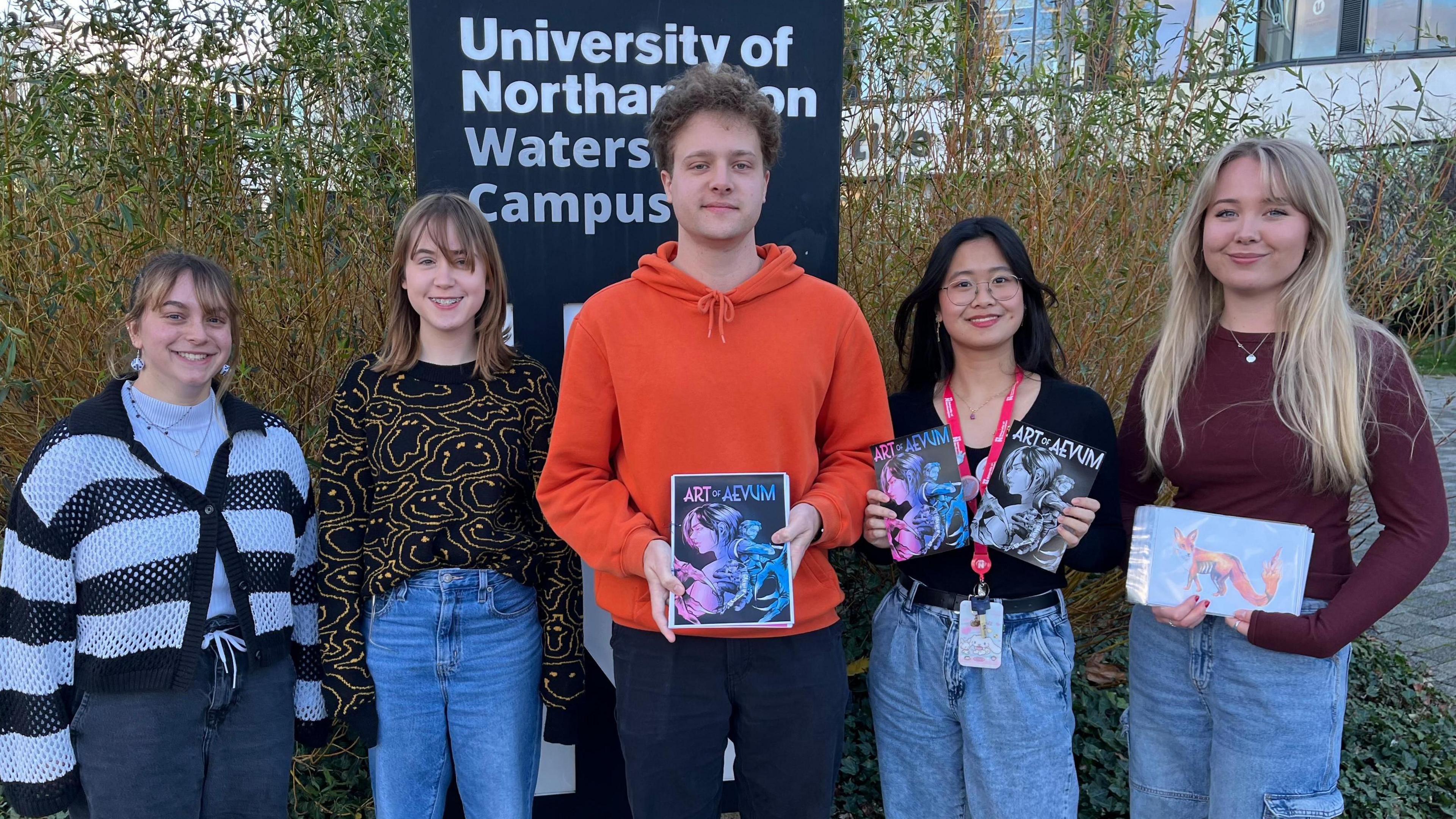 A male student in an orange jumper with two female students either side of him, holding their illustration display. All stood in front of a University of Northampton Waterside Campus sign.