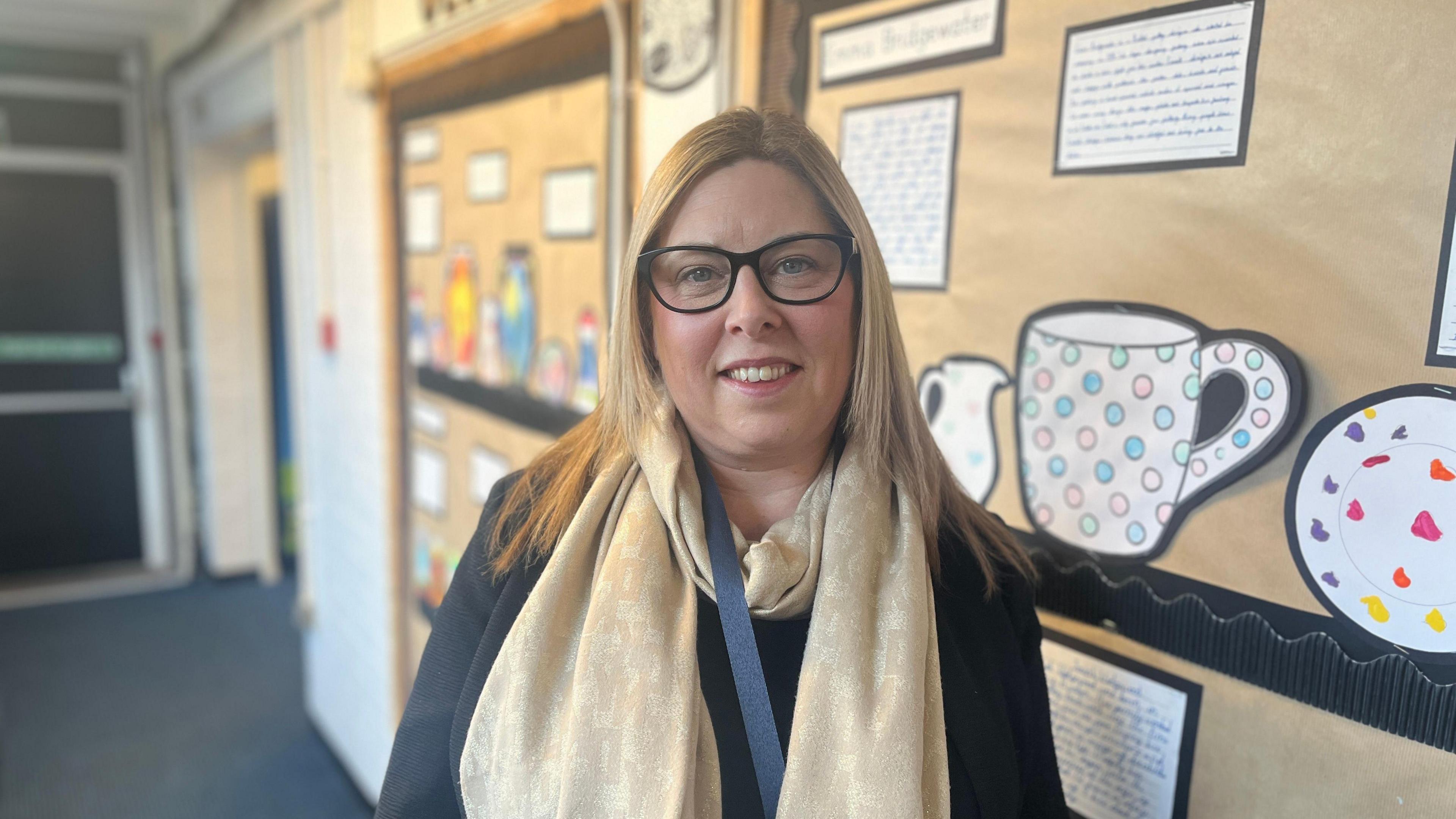 Head teacher Sarah Clowes is looking straight into the camera in the middle of the picture, in front of a wall display in a school corridor. She is smiling, has straight blonde hair and wears glasses, as well as a golden scarf.