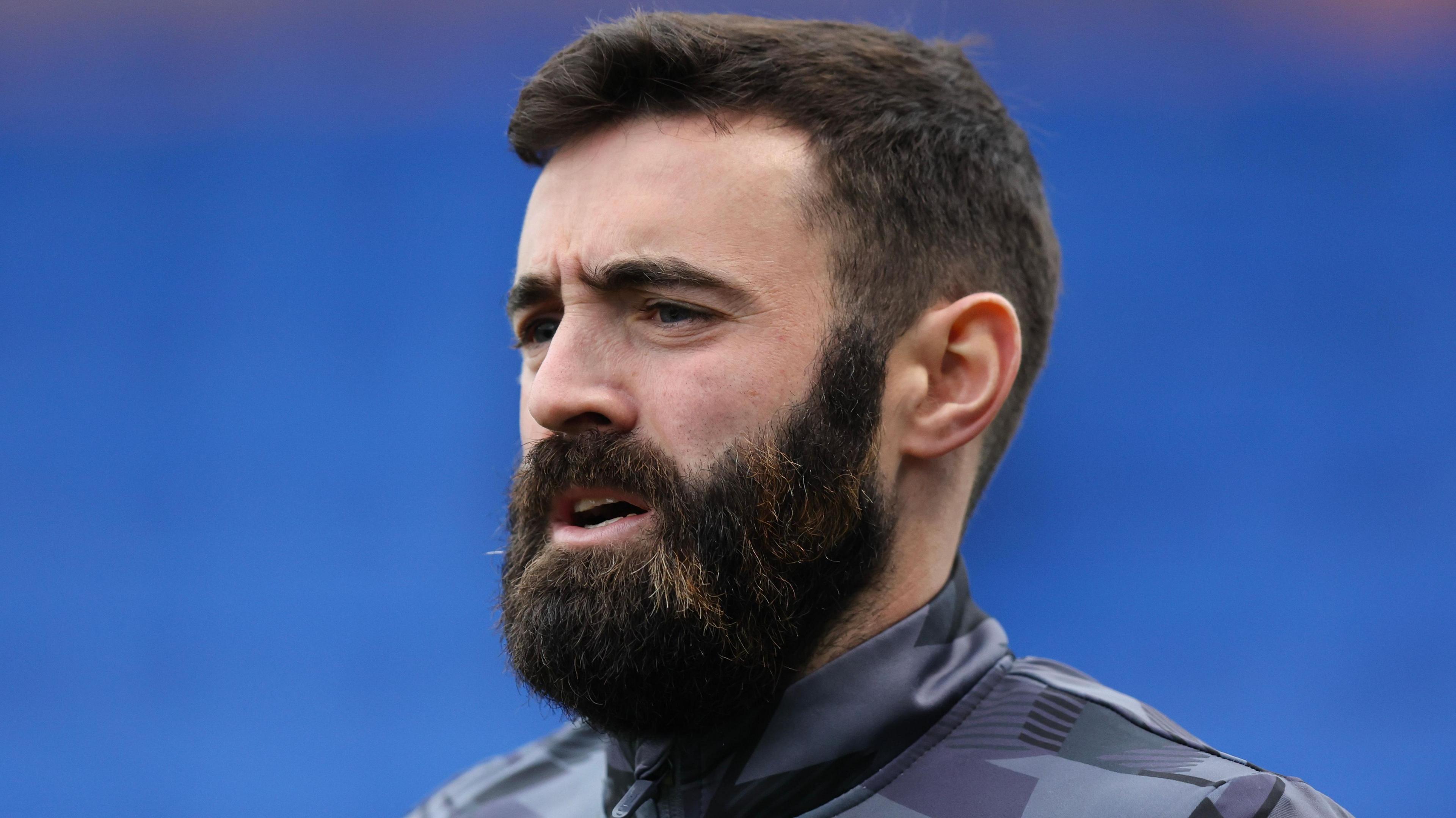 Jack Price, wearing a tracksuit and with a dark beard, against a blue background, looks forward