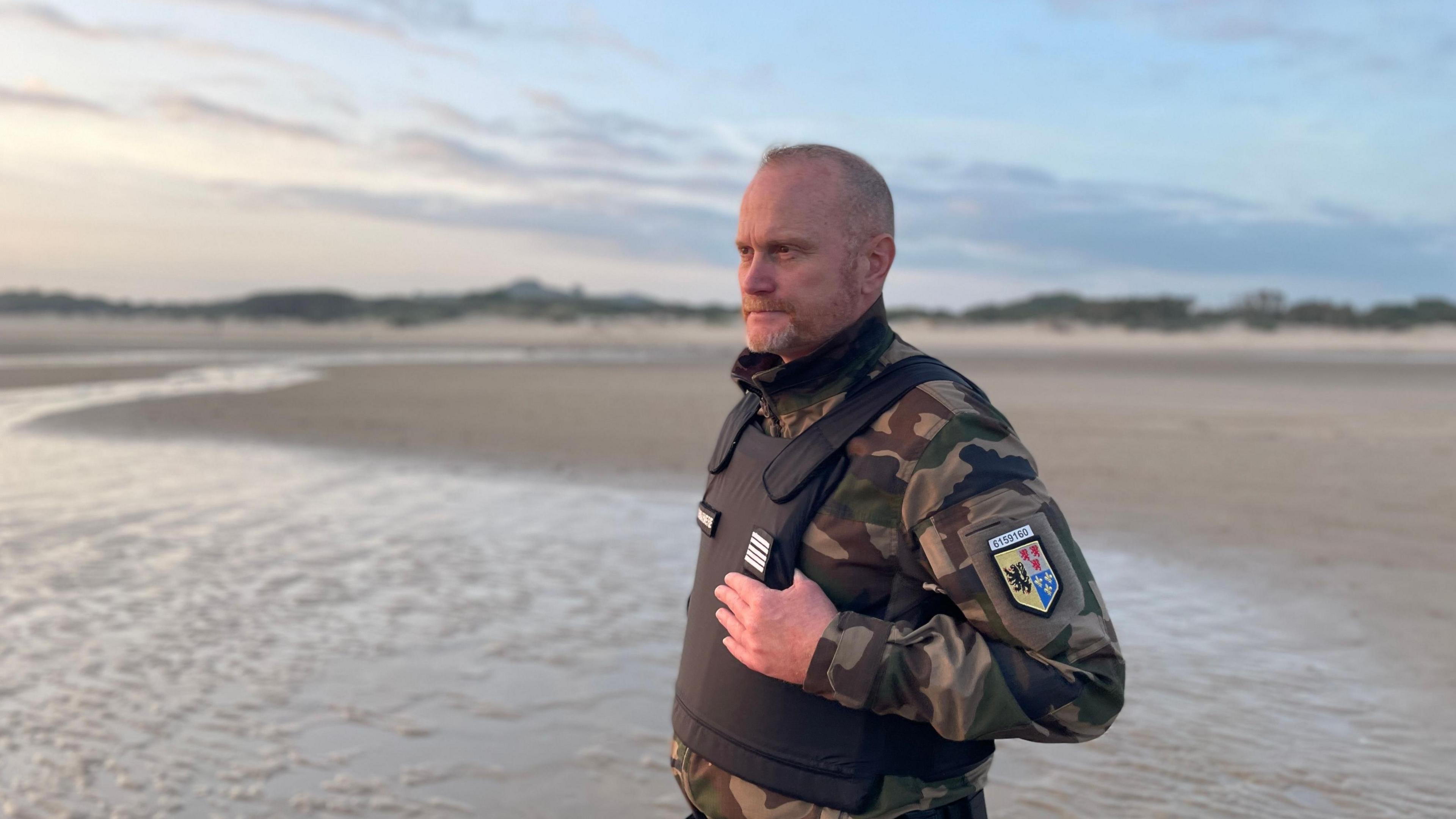 A man in his fifties dressed in police uniform, with a bullet-proof vest, stands on the sand of a beach looking out to see