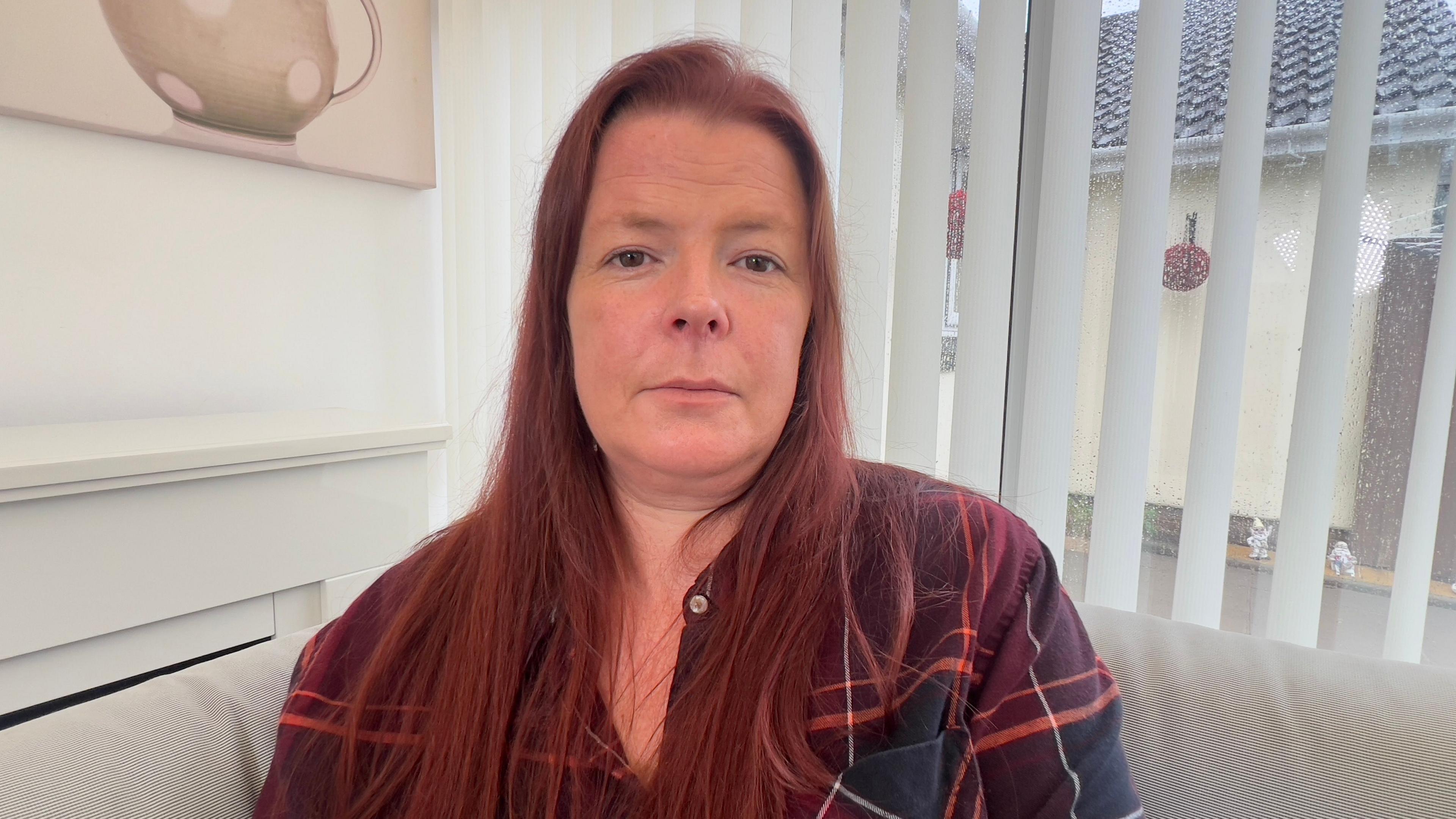 Rachel Kemble who has long red hair, sat on a cream corner sofa in front of windows with blinds on, and wearing a red tartan shirt