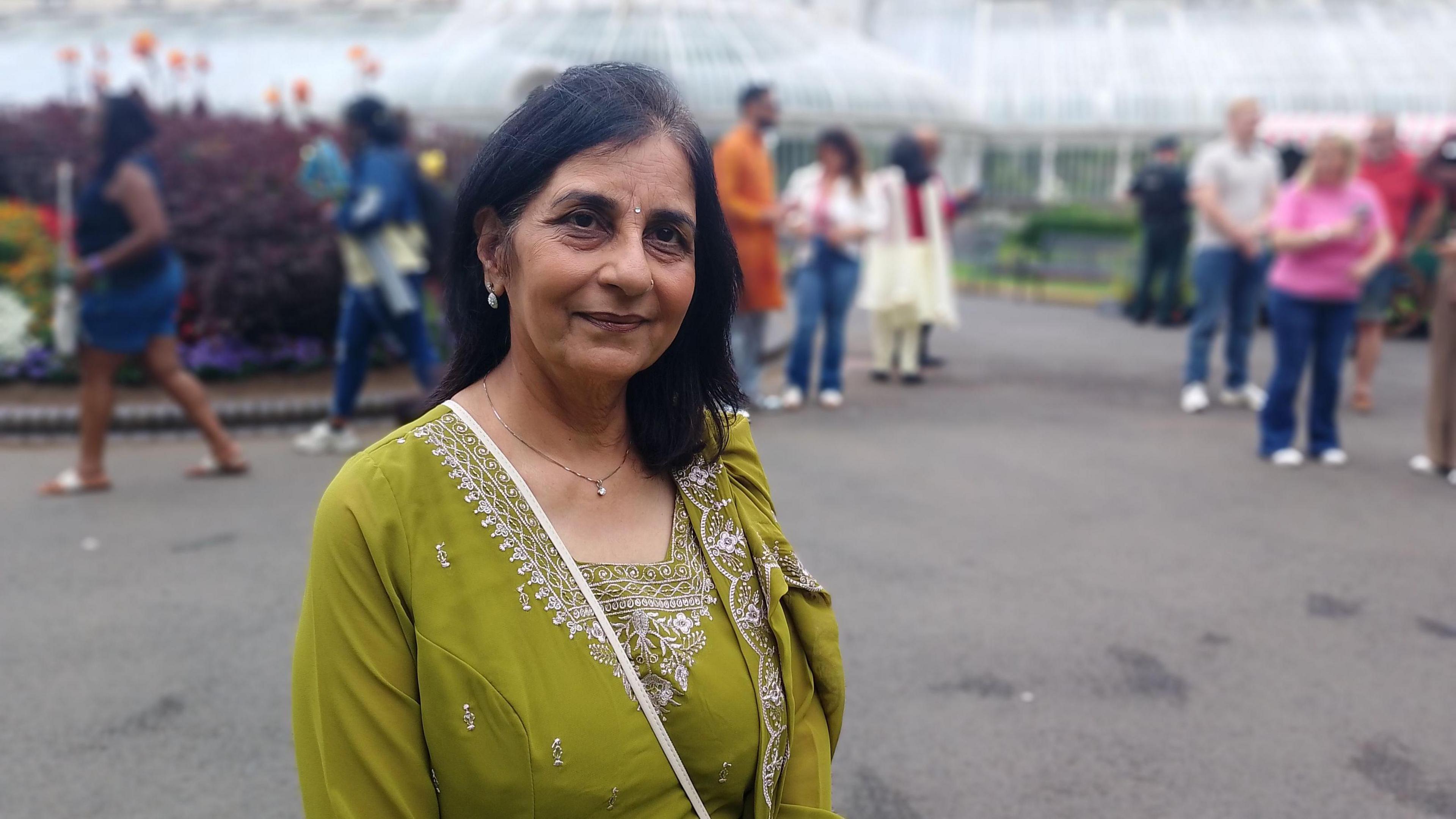 Dr Nisha Tandon is dressed in green. She has black hair. She's standing in the Botanic Gardens with a hothouse behind her. People are behind her in the background.