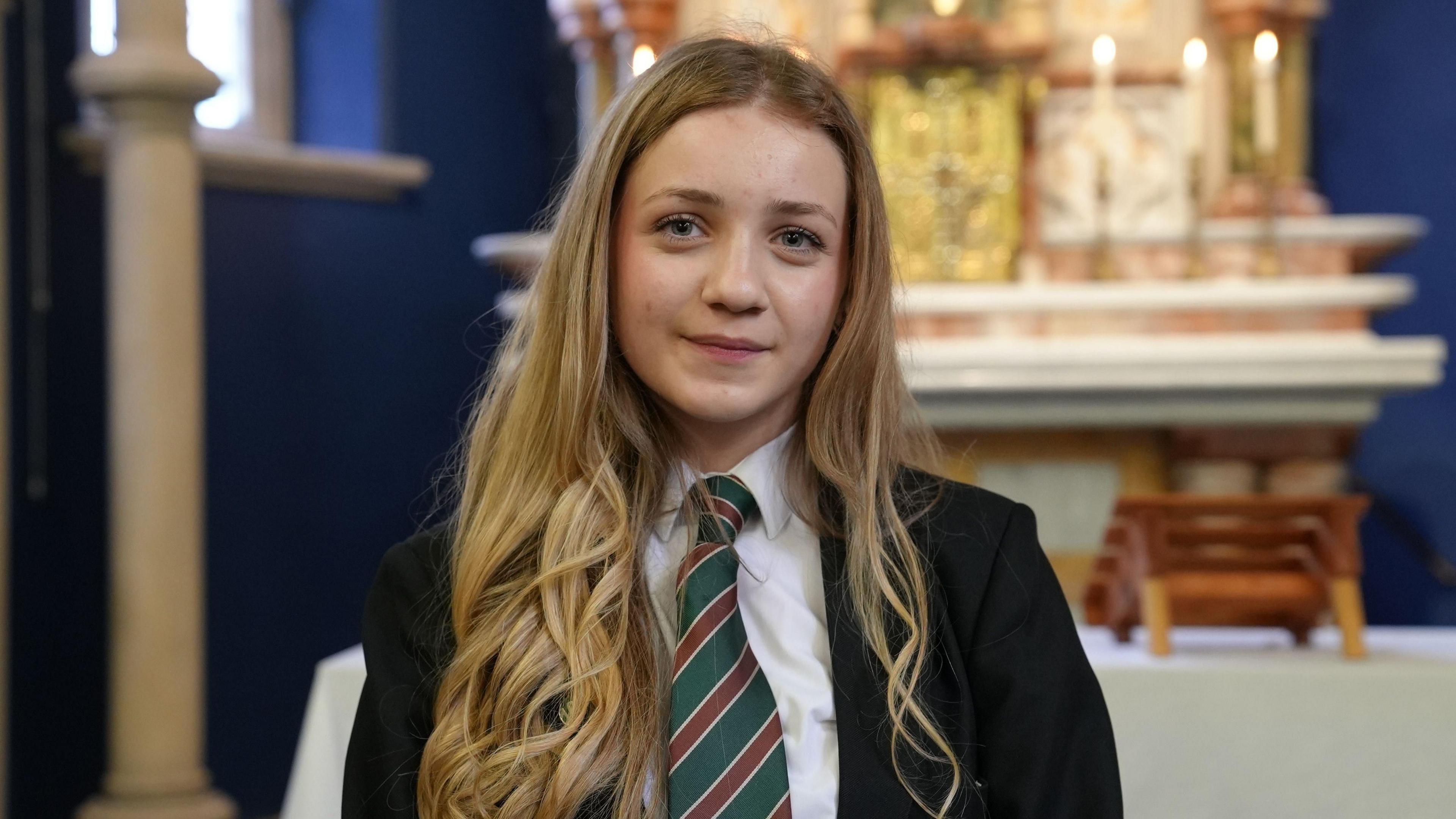 A schoolgirl in her uniform with long blonde hair standing in front of an altar in a church