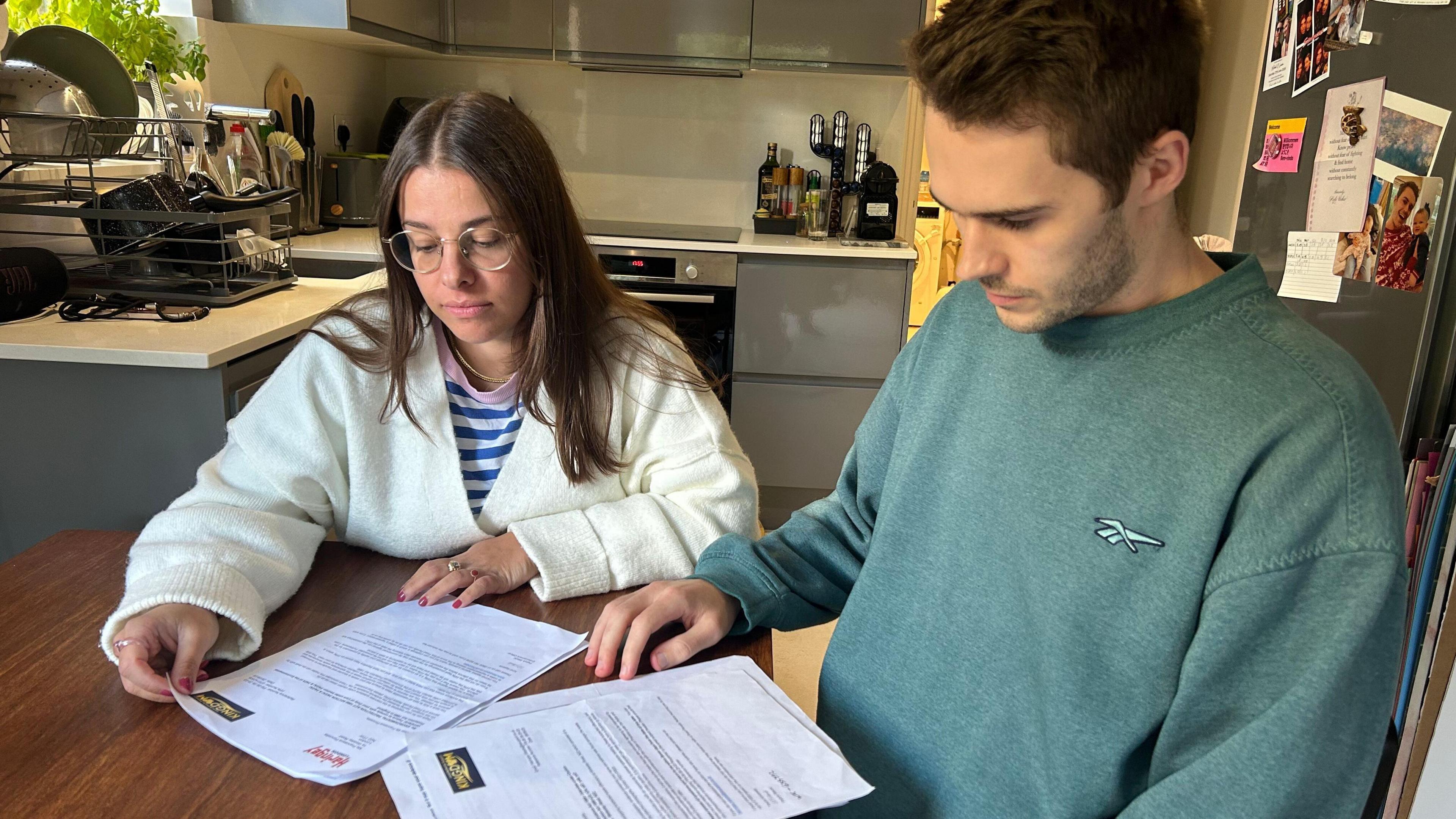 A young couple sit around a dining table in a modern kitchen looking mournfully at letters in front of them. 