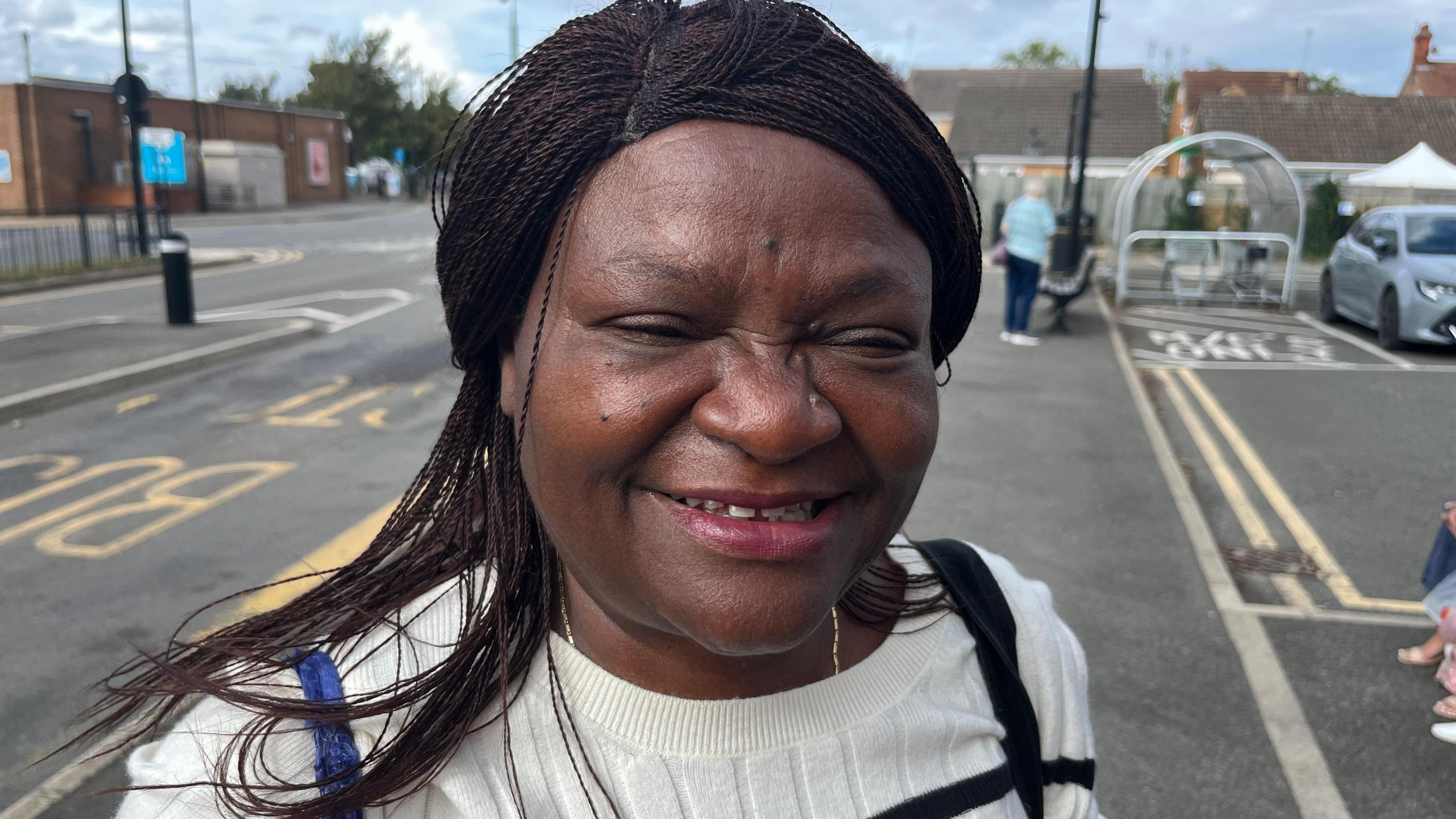 Ms Fredericks is standing at the bus stop and smiling. She has long dark hair and is wearing a gold chain and black and white horizontal striped top. In the road to her left is yellow painted letters that read 'Bus Stop'.