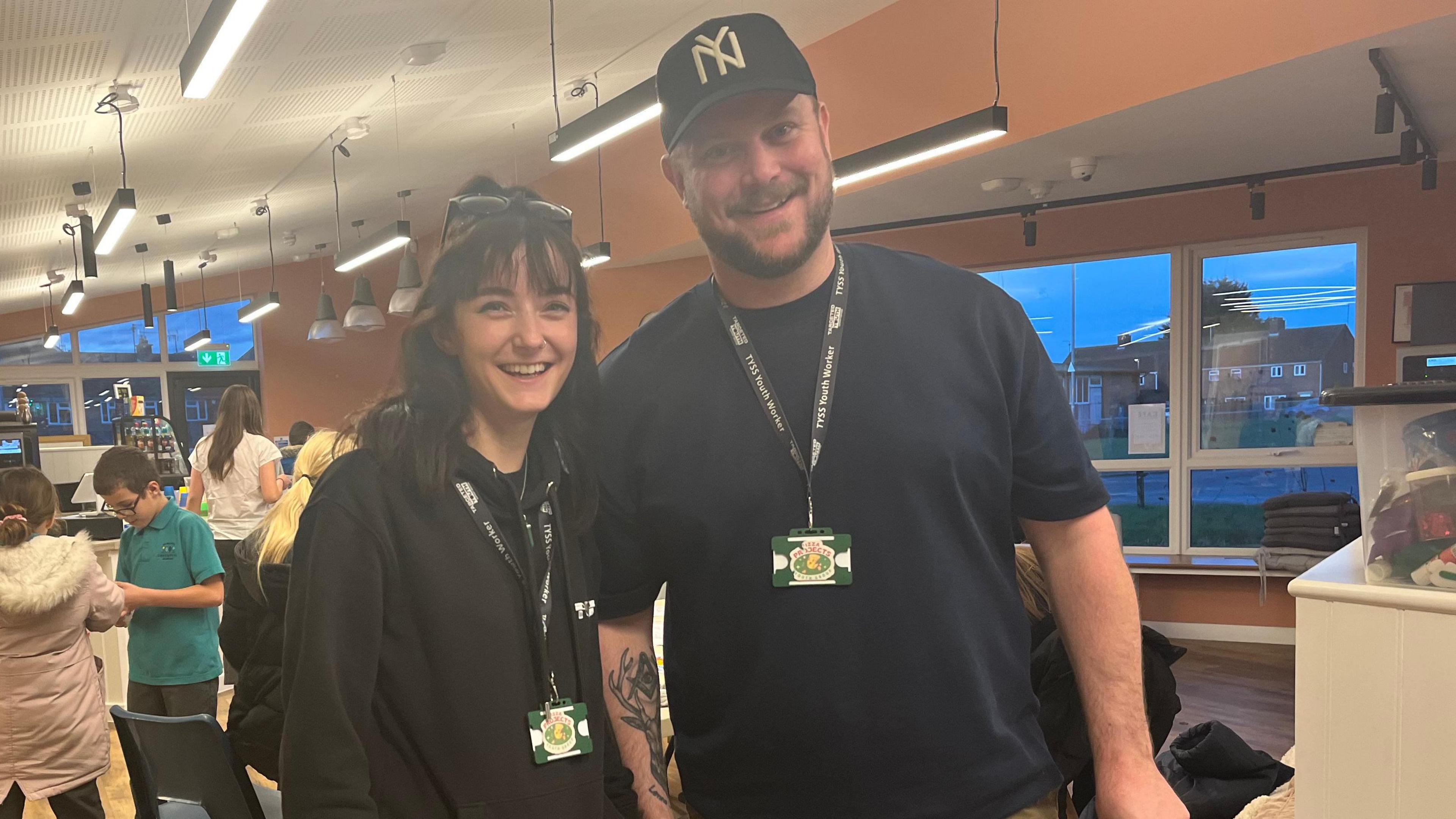 Youth workers Fiona Chaddock and Ben Griffin help run the Pizza Project. Fiona is dressed in black and has her glasses pushed up into her hair. Ben is wearing a black cap and a dark blue t shirt. He has a tattoo on his forearm. 