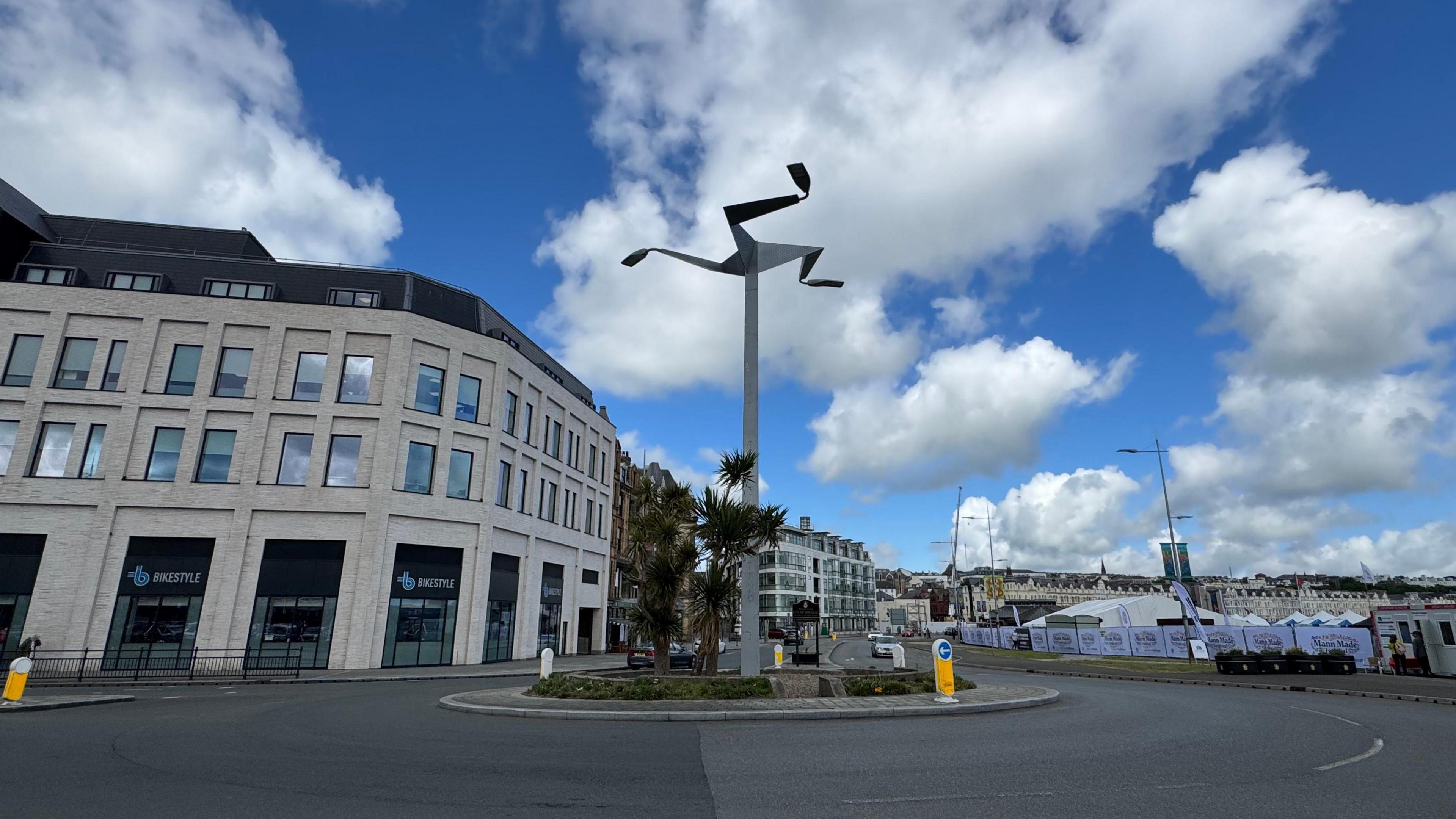 A tall silver column, with the three legs of mann, stands above a roundabout. There is a modern light building in the background and marquis tents on the other side.