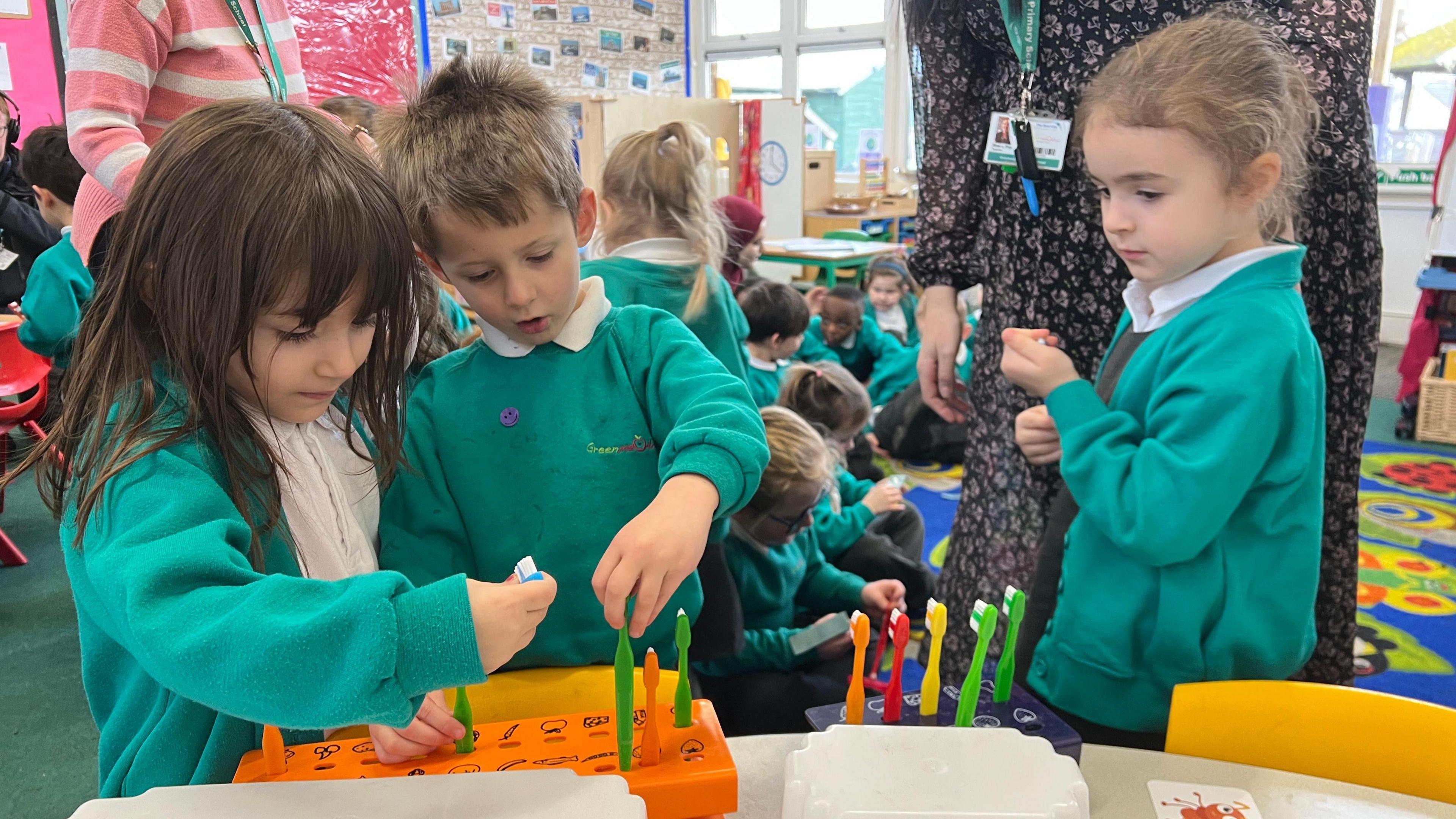 Three schoolchildren wearing teal uniforms examine and choose multicoloured tooth brushes in a school classroom. A number of children wearing the same uniform and adults wearing cardigans and dresses appear in the background.