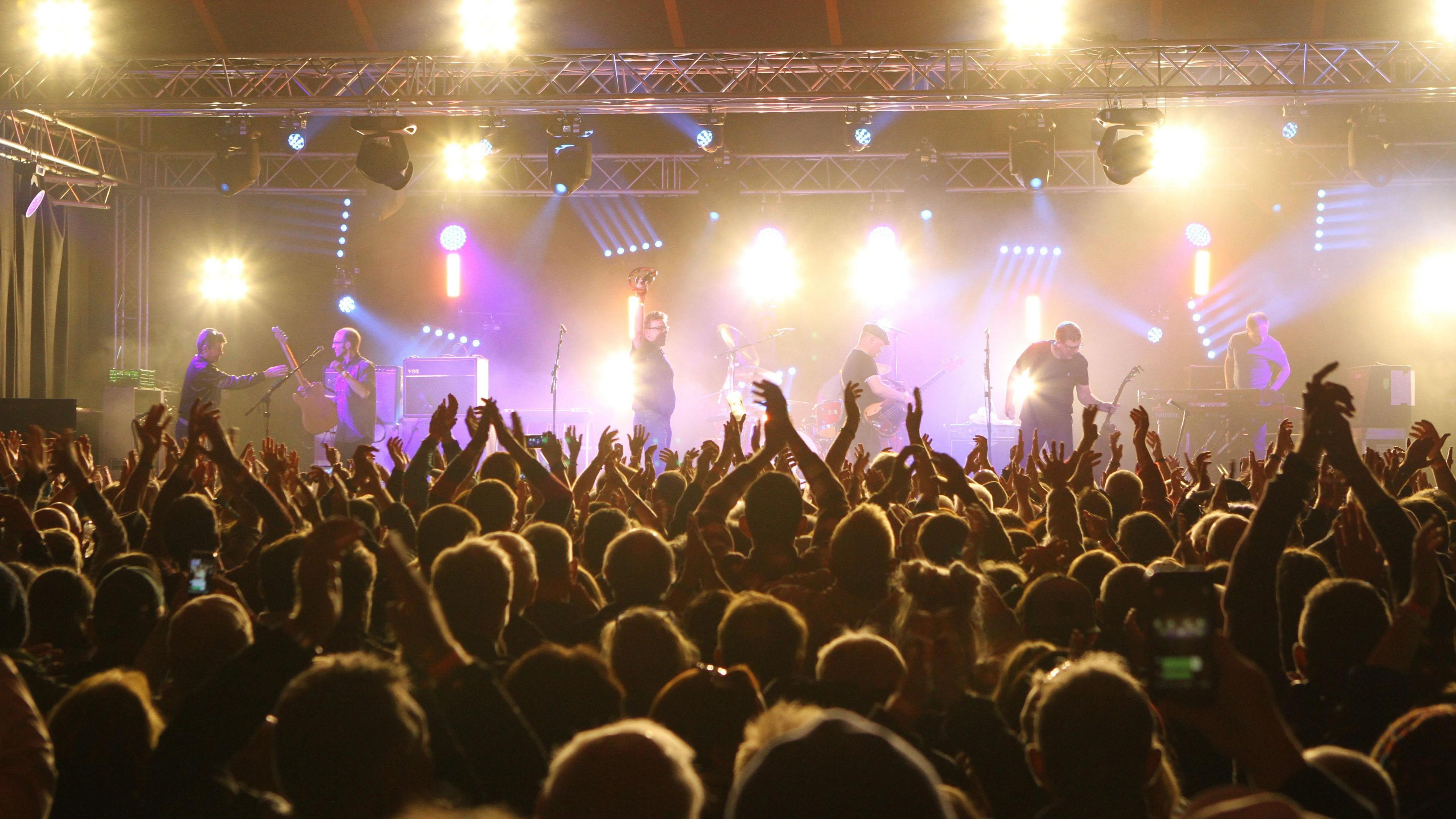 A crowd at the Towersey Festival with a band on the stage. There are bright stage lights. One of the band members has one arm in the air, holding a tambourine.