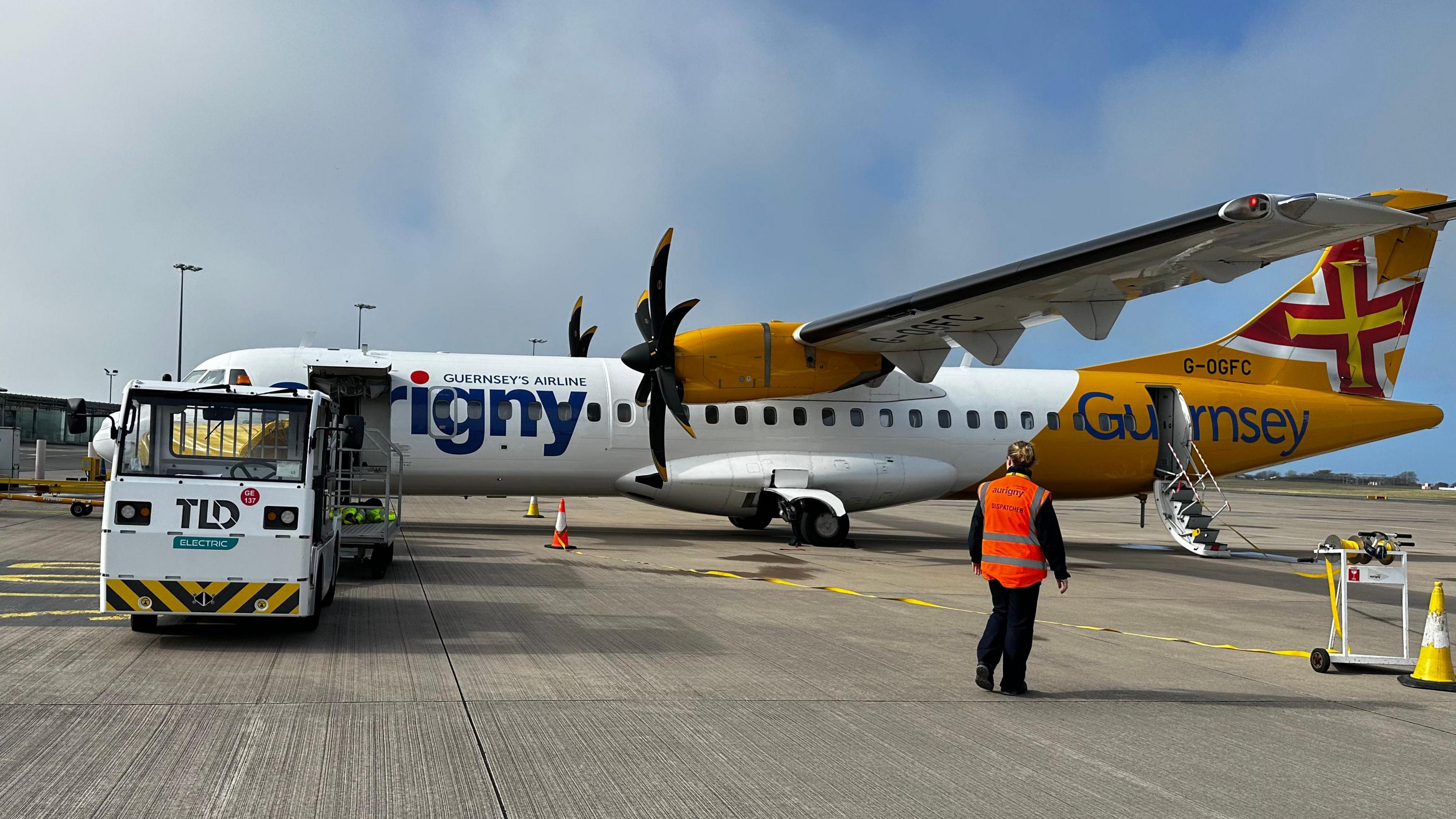Ground crew workers preparing an Aurigny plane. The aircraft is yellow and white with two propellers - one on each wing. A worker in an orange high-vis jacket is walking along the tarmac.