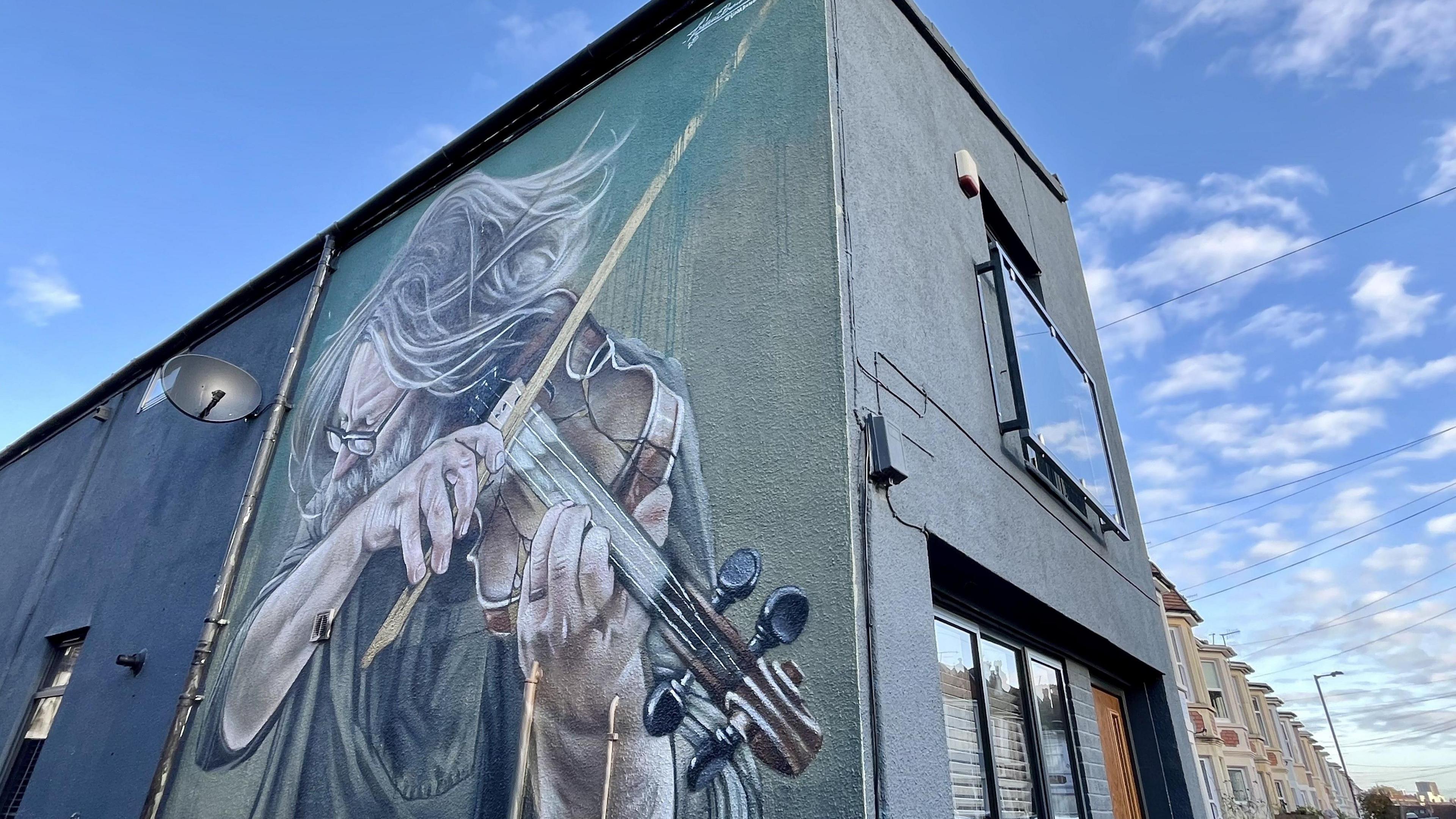 A large street mural is visible on a side street in Southville in Bristol. The exterior of the house is a dark green colour and the mural is of an elderly man with white hair and a white beard playing the violin