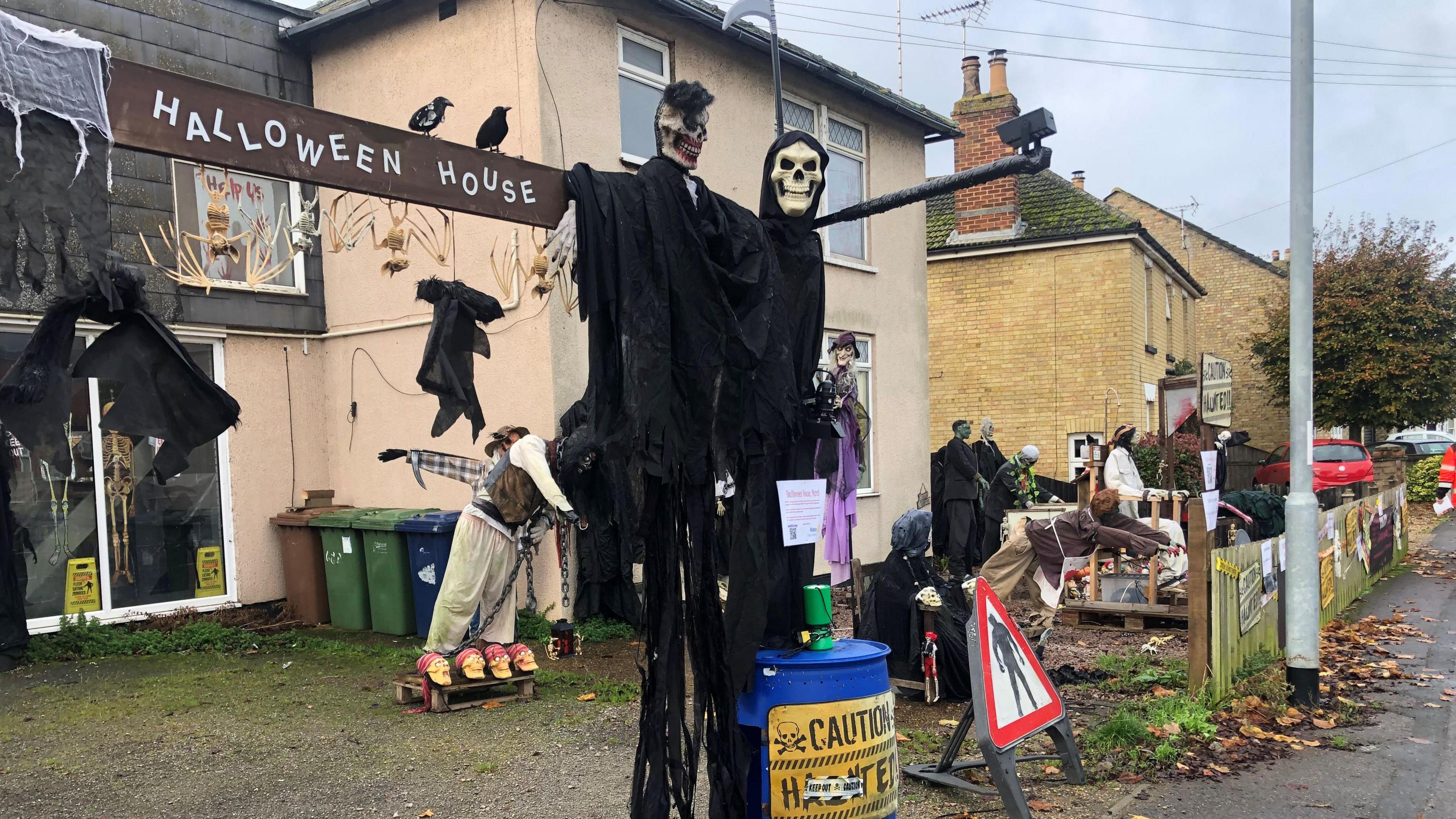 A residential road with a house that has a host of horror dummies and mannequins, there is a tall sign, about 3m high saying, 'Halloween House'. The two-storey house is cream coloured, has patio sliding doors to the left, and bedroom windows visible on the second floor.