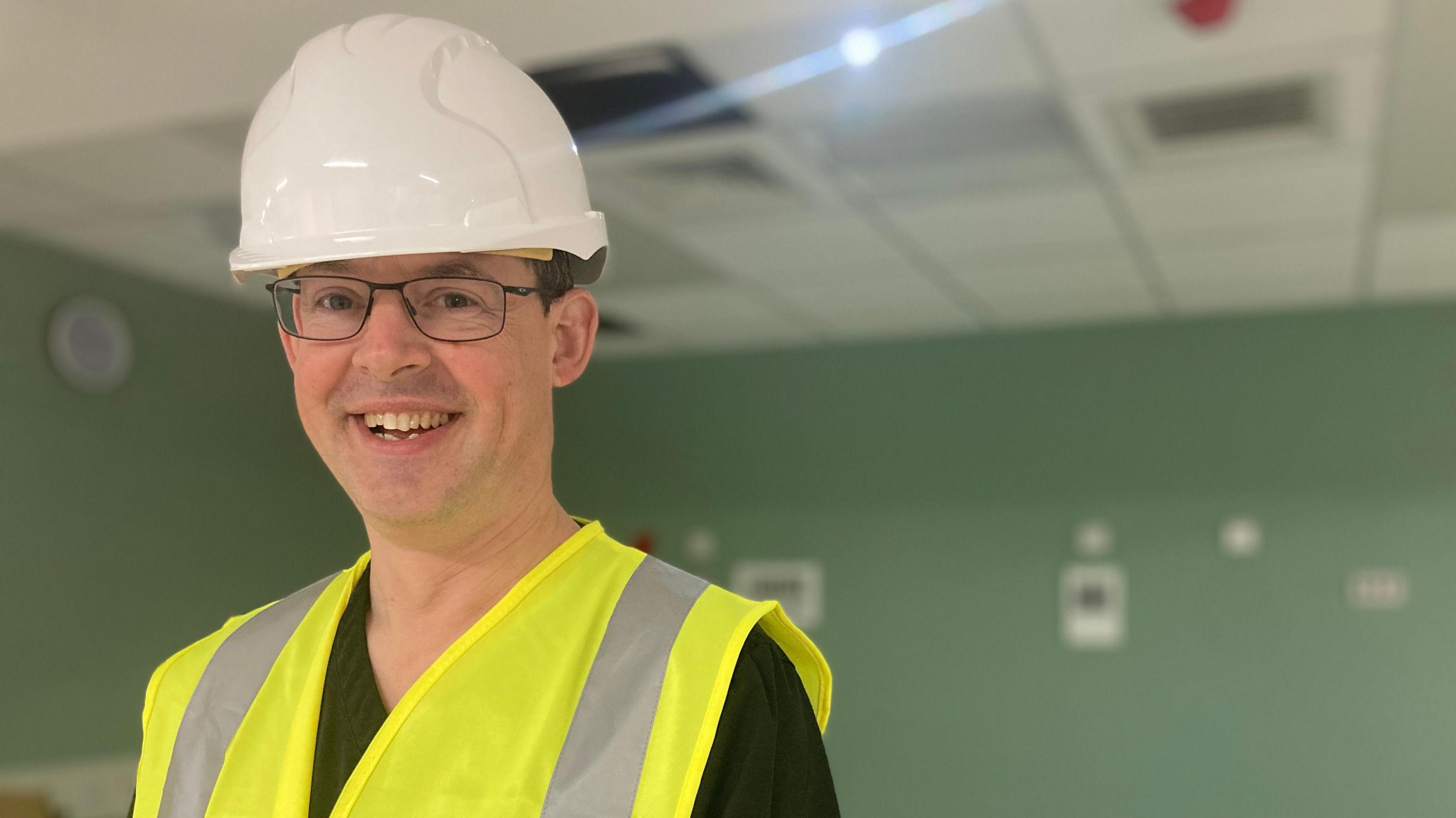 A man in a white hard hat and wearing a hi-vis vest smiles towards the camera. He is in room with green walls and a white suspended ceiling