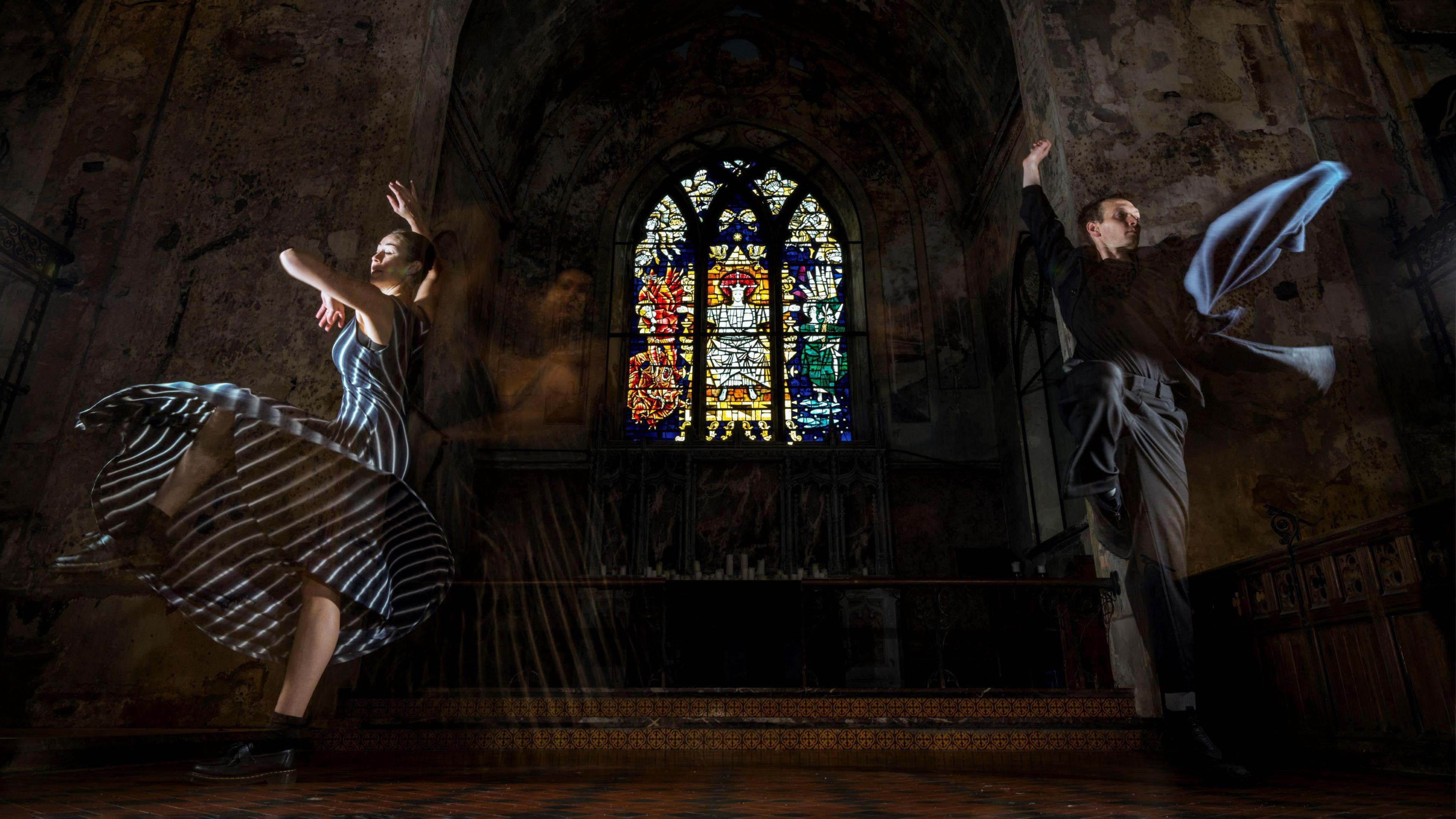 Two performers from Bristol dance group Impermanence rehearse inside venue The Mount Without. One is a woman wearing a long blue and white striped dress and the other is a man in black robes. They are both in the air as they perform dance moves