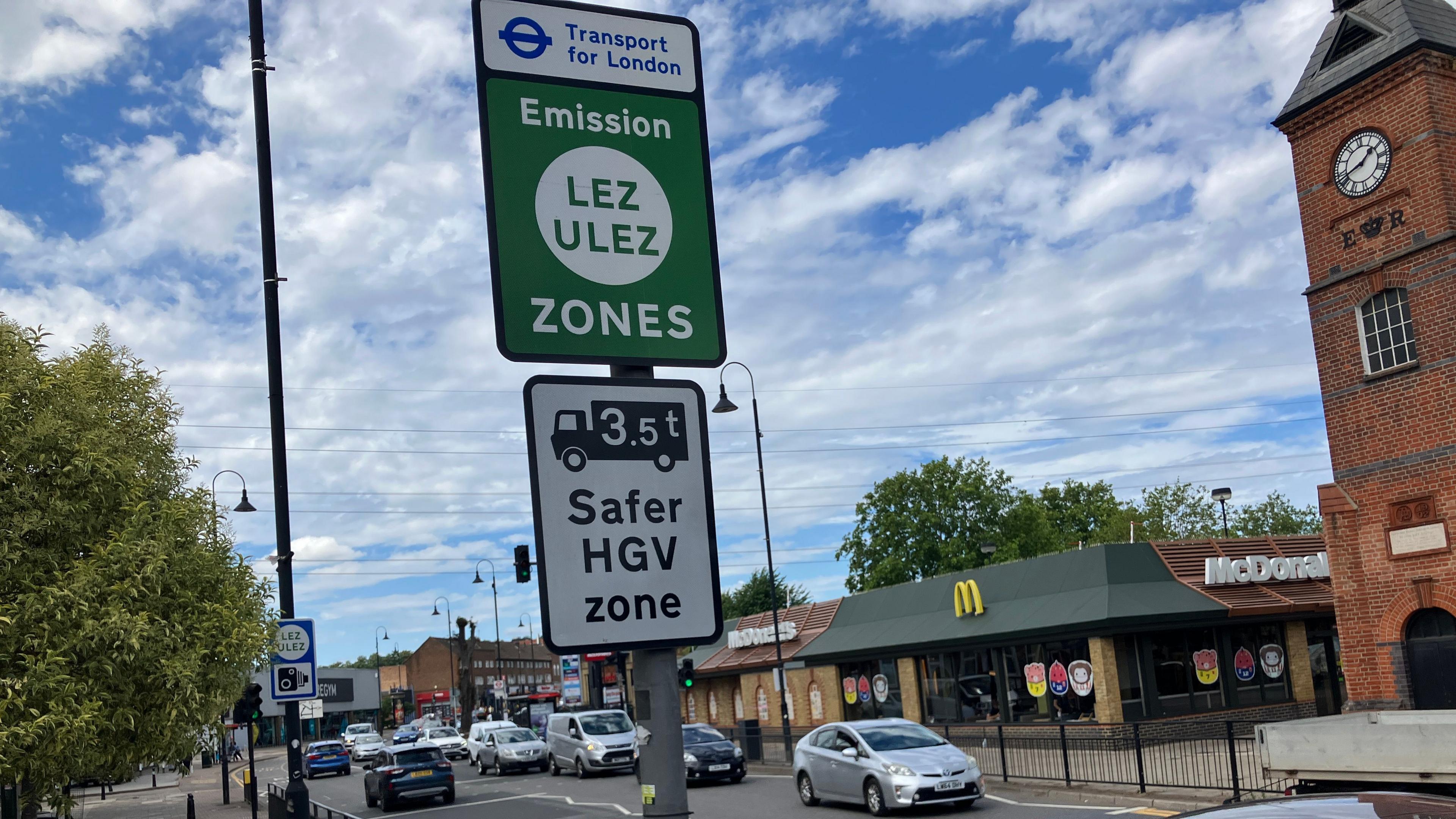 A Transport for London roadside sign marking the LEZ and ULEZ zones is mounted on a pole beside a busy road. Cars are travelling through a junction and a McDonald's restaurant and brick clock tower can be seen in the background under a partly cloudy sky.