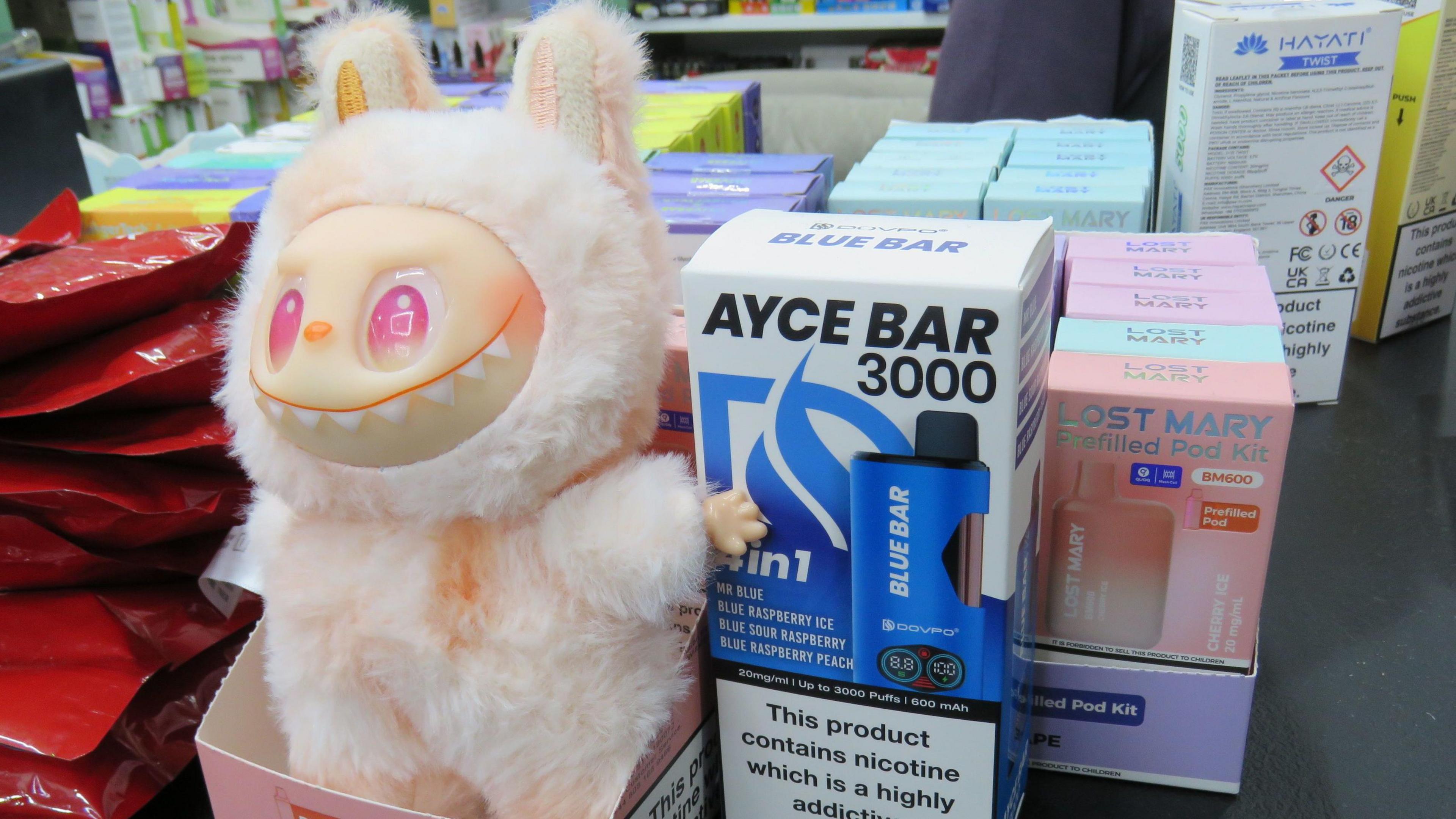 A pink and white labubu (a plush monster toy with exaggerated facial features and jagged grin) stands next to boxes of vapes on a shop counter.  