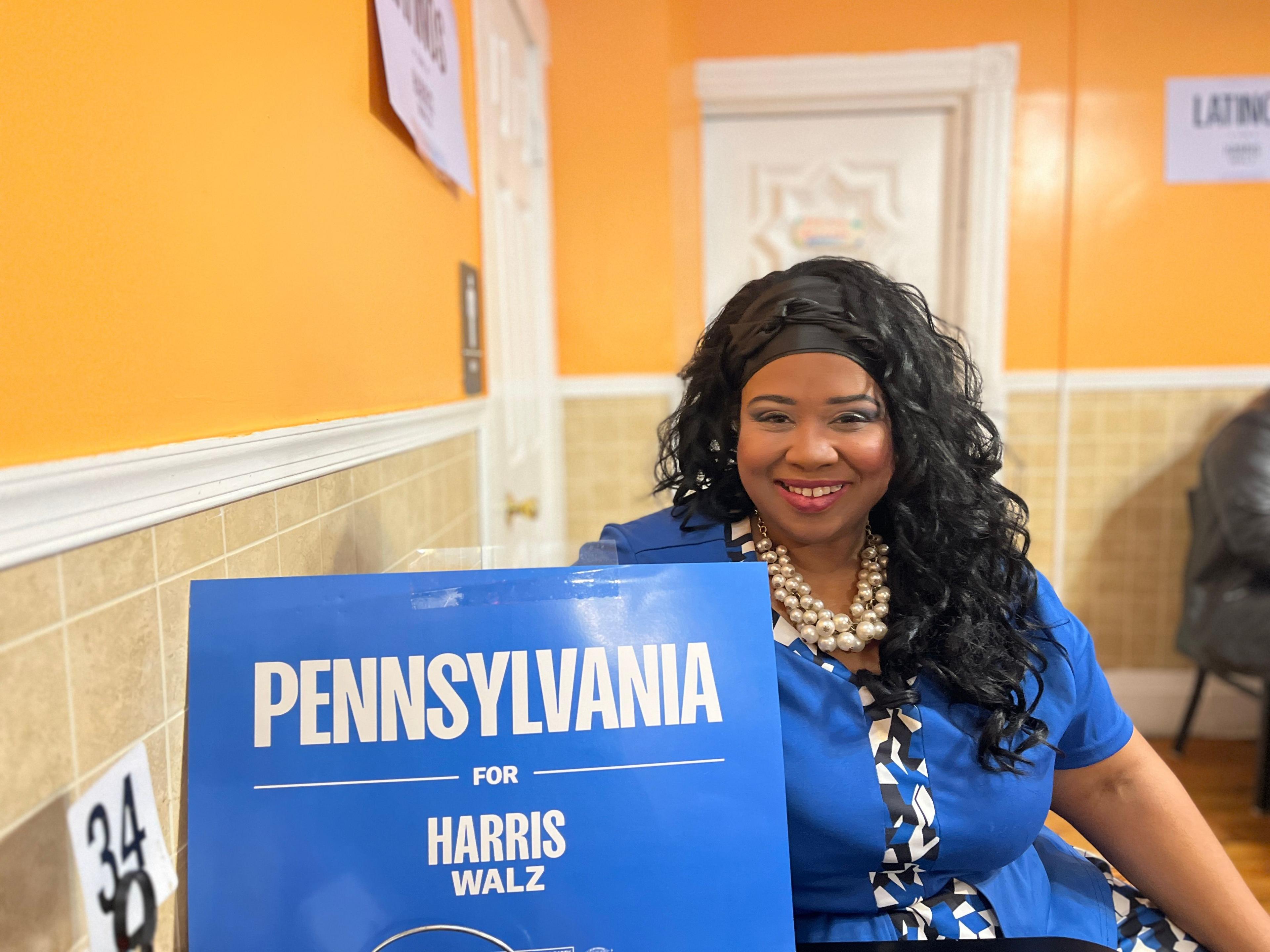 Kari Holmes, wearing a blue shirt, smiles while holding a Pennsylvania for Harris Walz sign