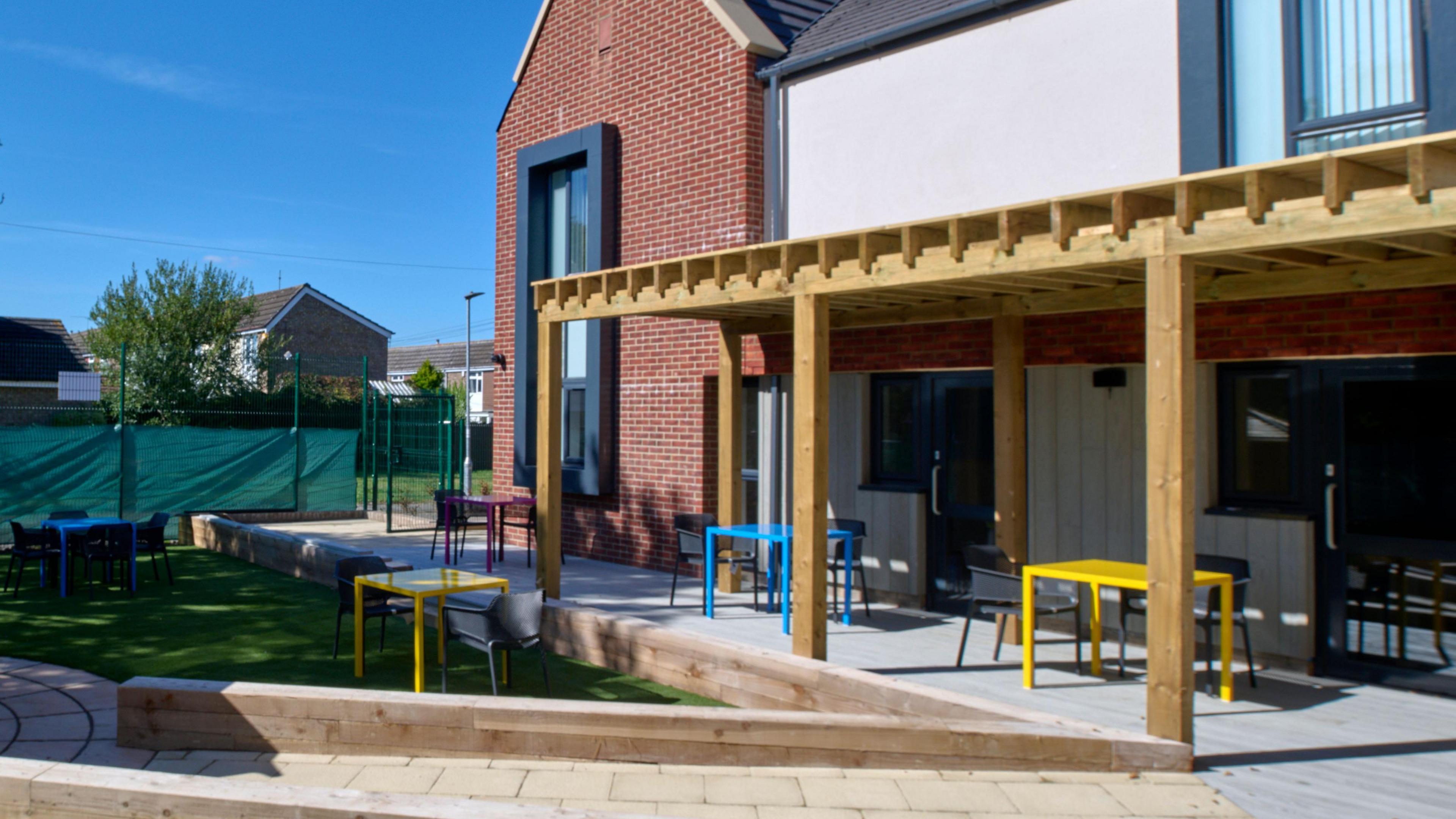 The outside of a modern building with large glass windows and a patio area with brightly coloured chairs and tables leading on to the garden.