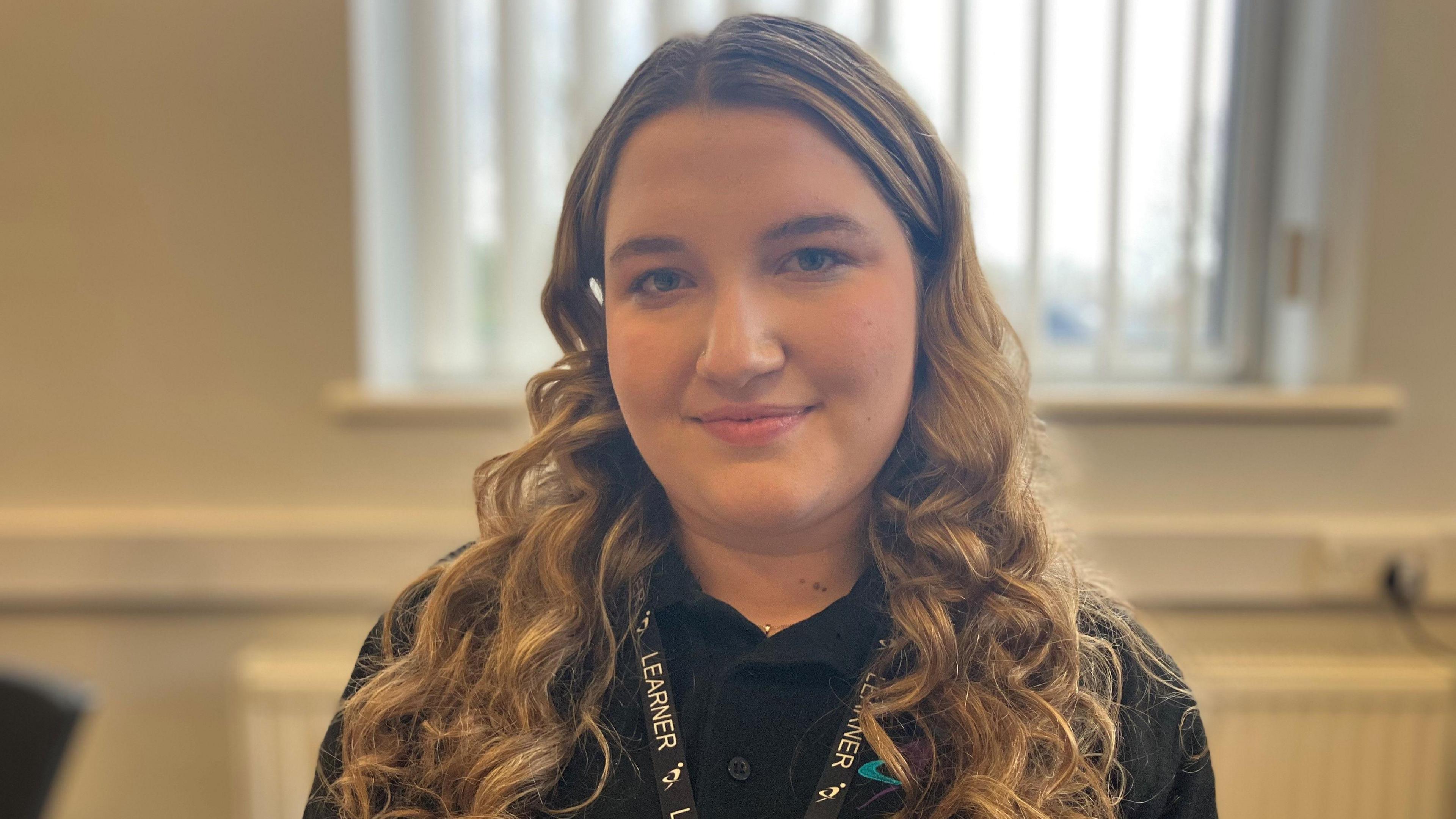 Roma, who has fair, long curly hair and his wearing an ID lanyard over a black top in a classroom at Dudley College