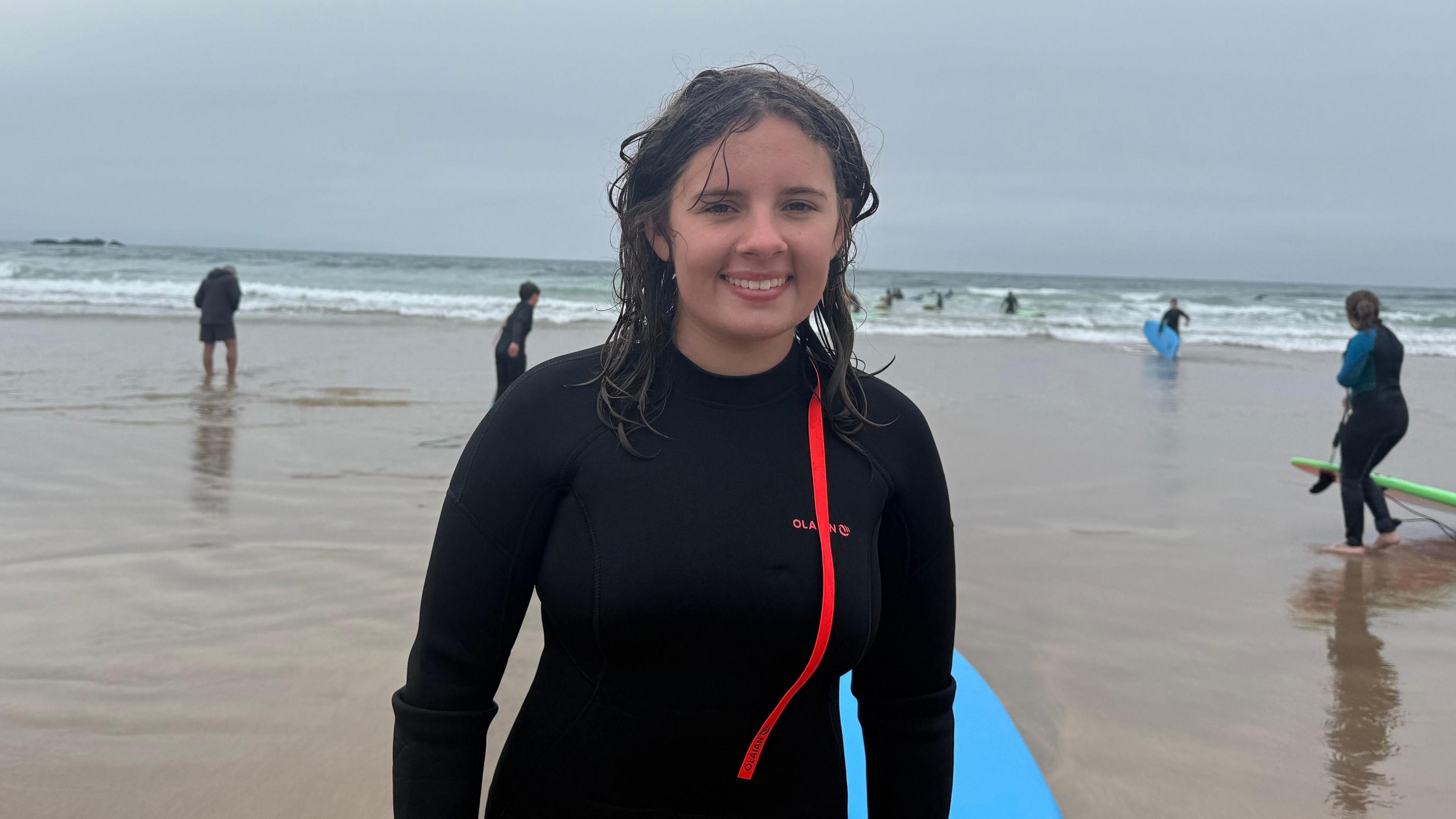 Eve Curry. She has long brown hair, wearing a black wetsuit with orange stripe. She has just come out of the water and is standing next to a blue surfboard.