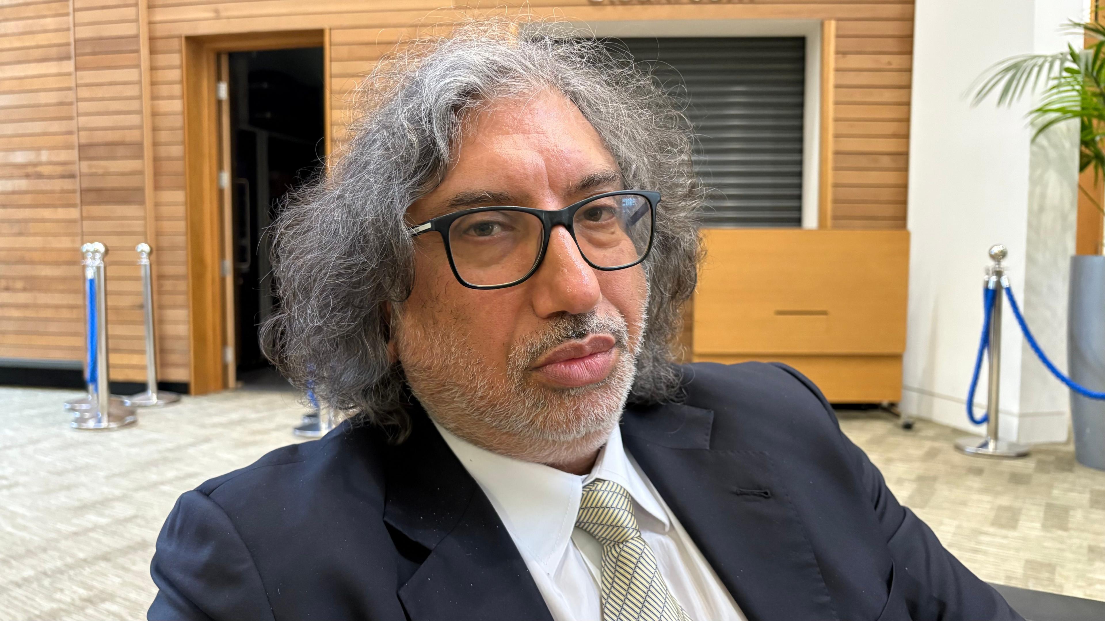 Niknam Hussain with long grey hair, slight beard and dark-rimmed glasses wearing a black jacket, white shirt and pale green tie. He is sitting back in a chair with a serving hatch set into a wooden wall behind him.