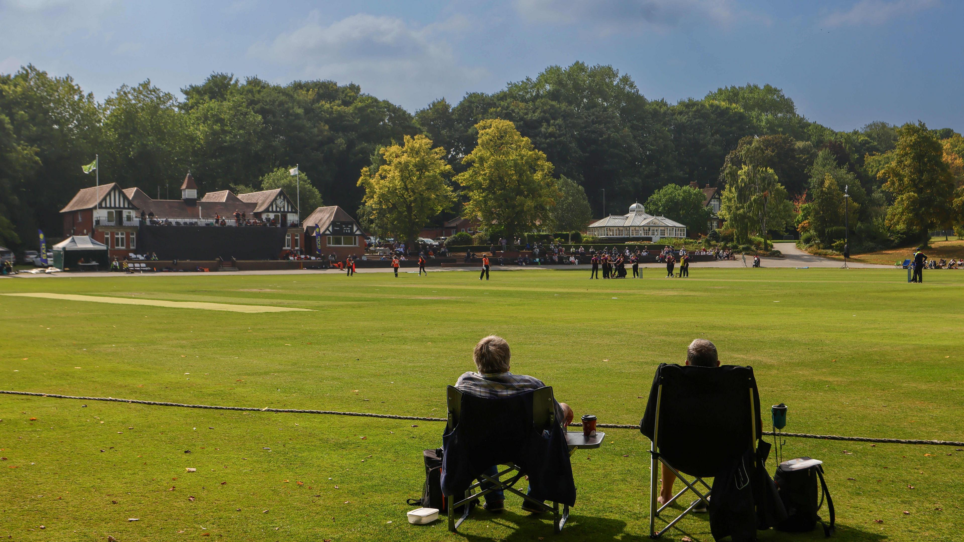 Two spectators sit on camper chairs with their back to the camera as they watch a Blaze women's cricket game at Chesterfield Cricket Club. The pavillion is on the left of the foreground and there are lots of trees in the backdrop 