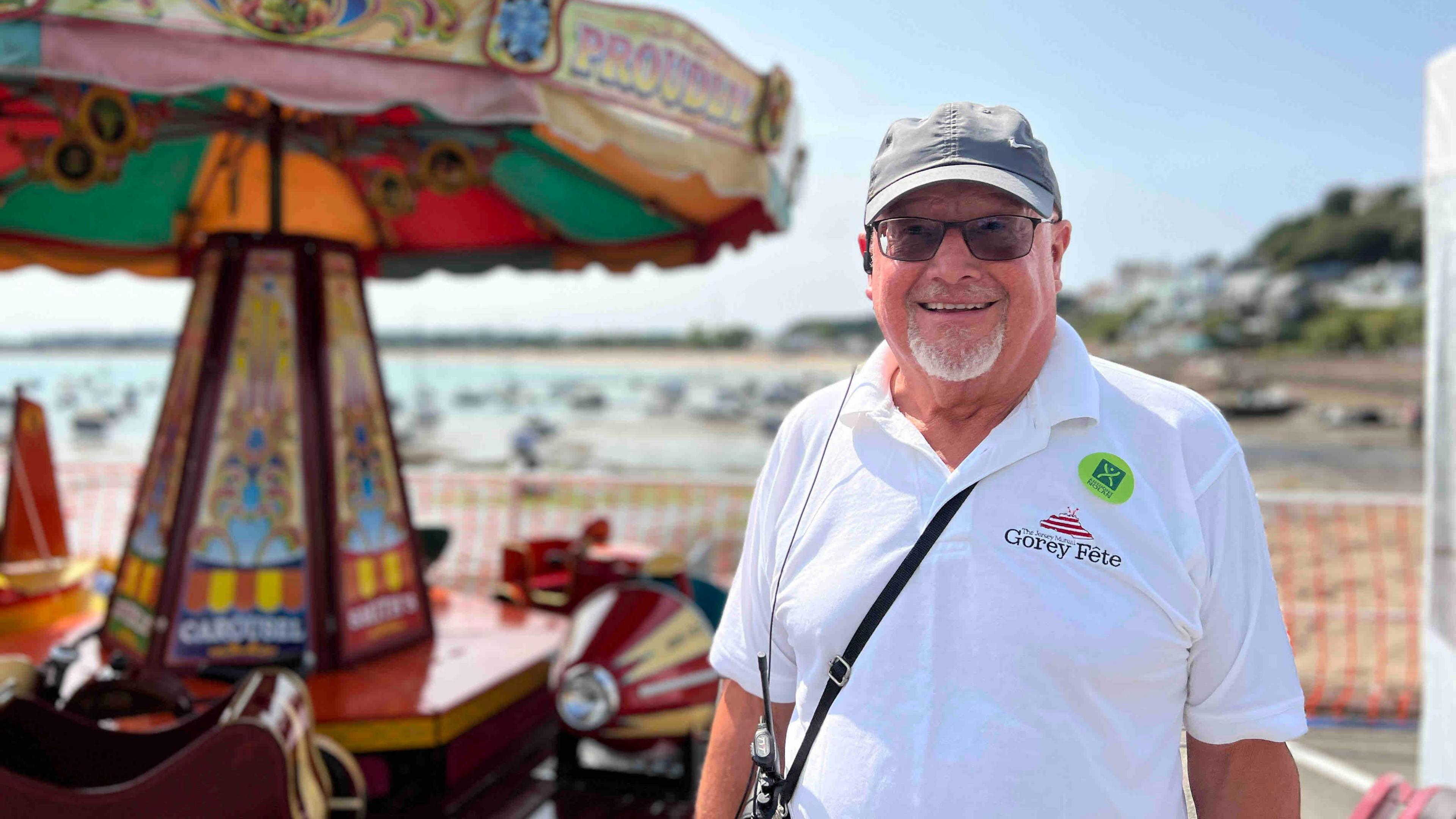 Lester Richardson is pictured in a white polo shirt. Mr Richardson is wearing a cap and sunglasses and he is smiling widely. There is a carousel behind him and a beach is also visible in the background.
