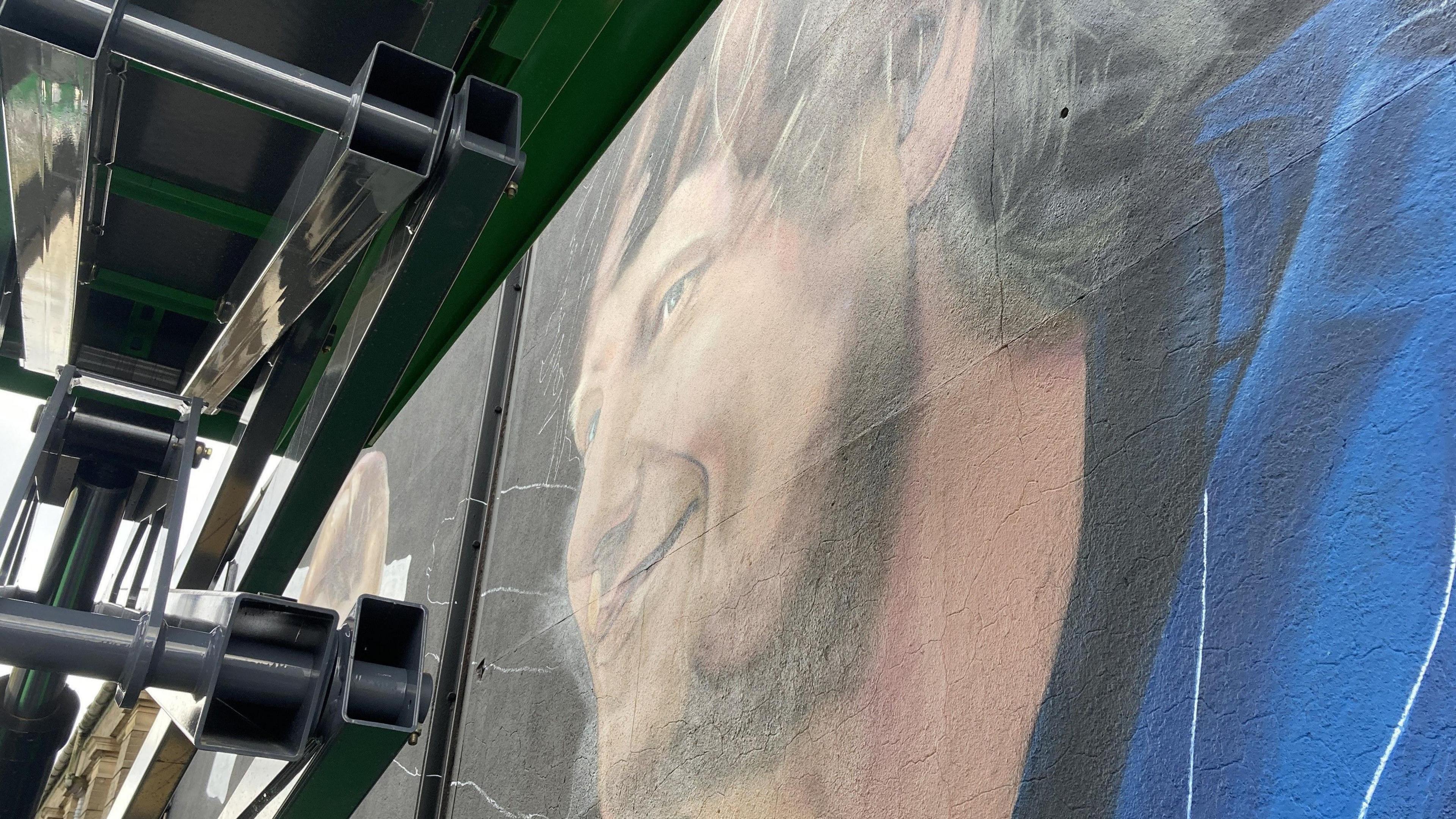 A closer view of the mural's older version of Sheeran. It depicts him smiling with ginger hair while wearing a blue Ipswich Town shirt. A scissor lift can be seen above the camera close to the mural wall. 