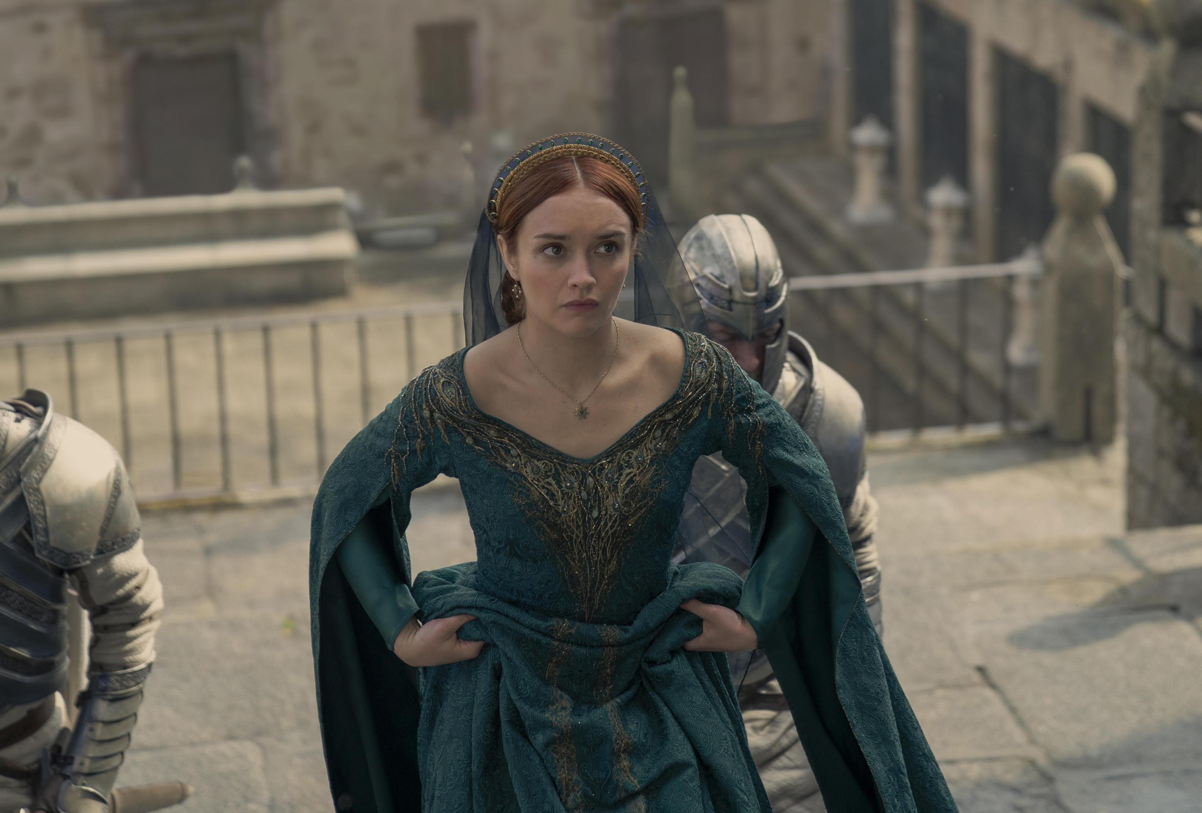 Red haired actress in green, embellished gown, with men in suits of armour, walking up steps in ancient looking location