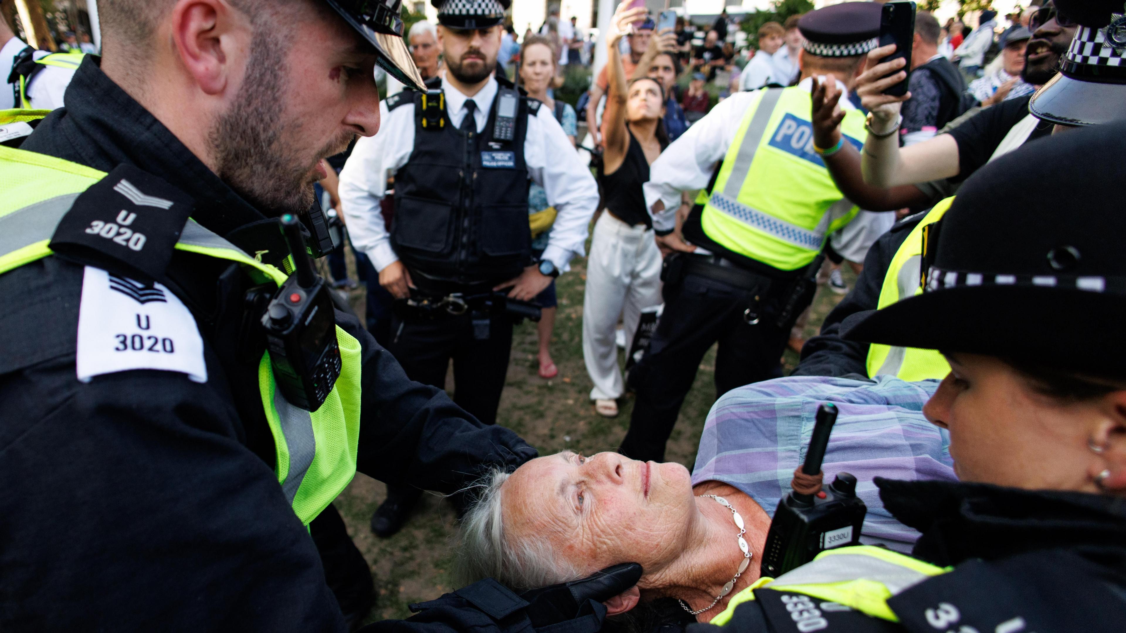 A woman with grey hair and wearing a necklace and checked shirt is carried away by officers, one of whom is holding her head as she lies still. In the background more officers and people taking pictures can be seen