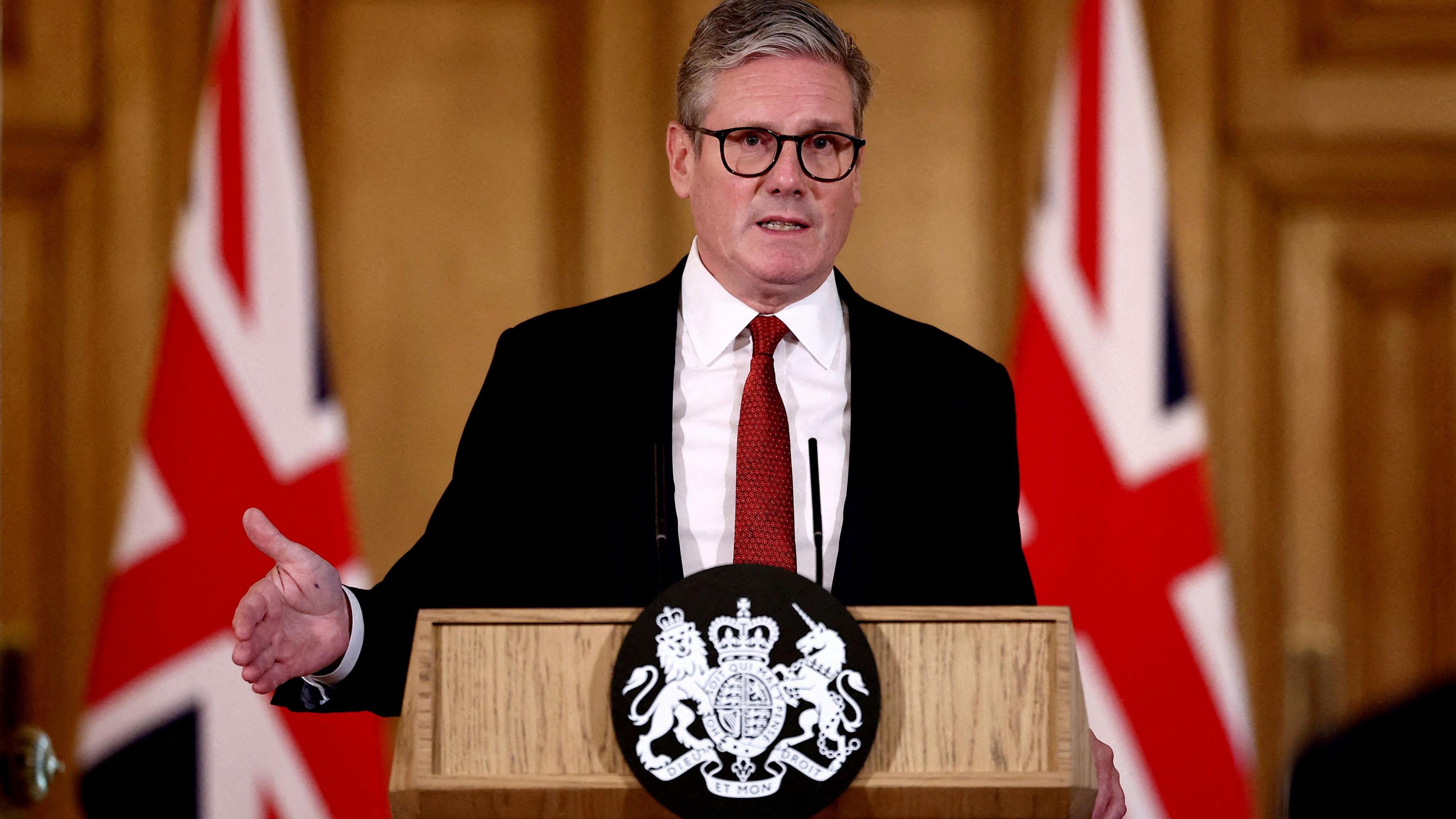 PM Sir Keir Starmer at a press conference with two flags of Great Britain behind him and wearing a black suit with a white shirt and red tie.