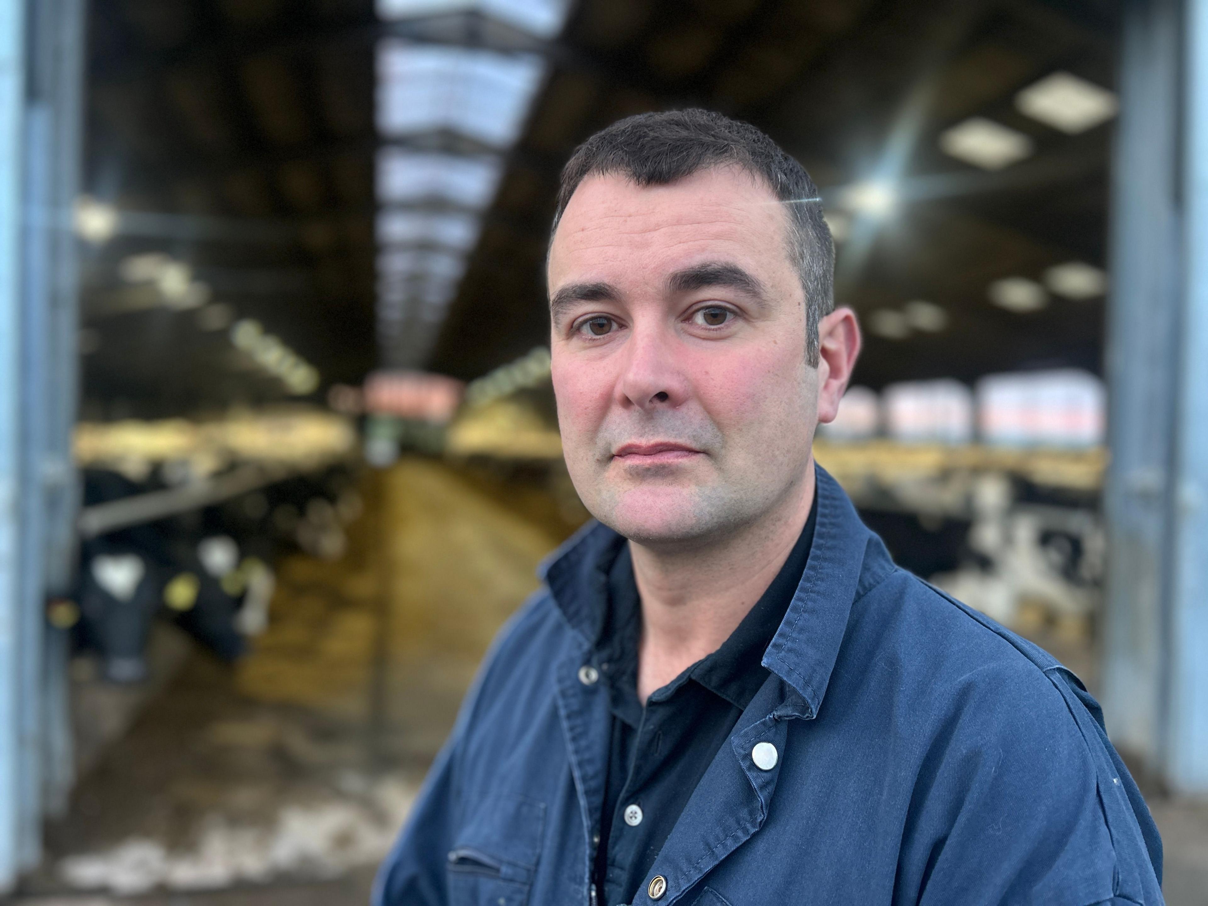David McMiken wearing a blue boiler suit and standing in front of his cow shed