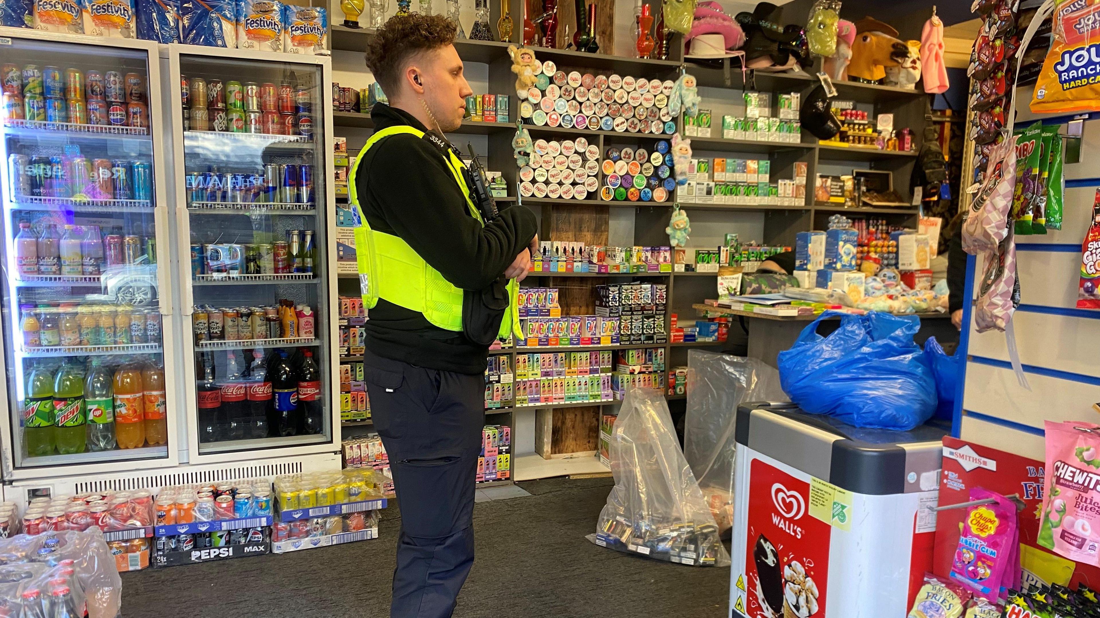 A police officer stands in a shop which is full of drinks, sweets and vapes.