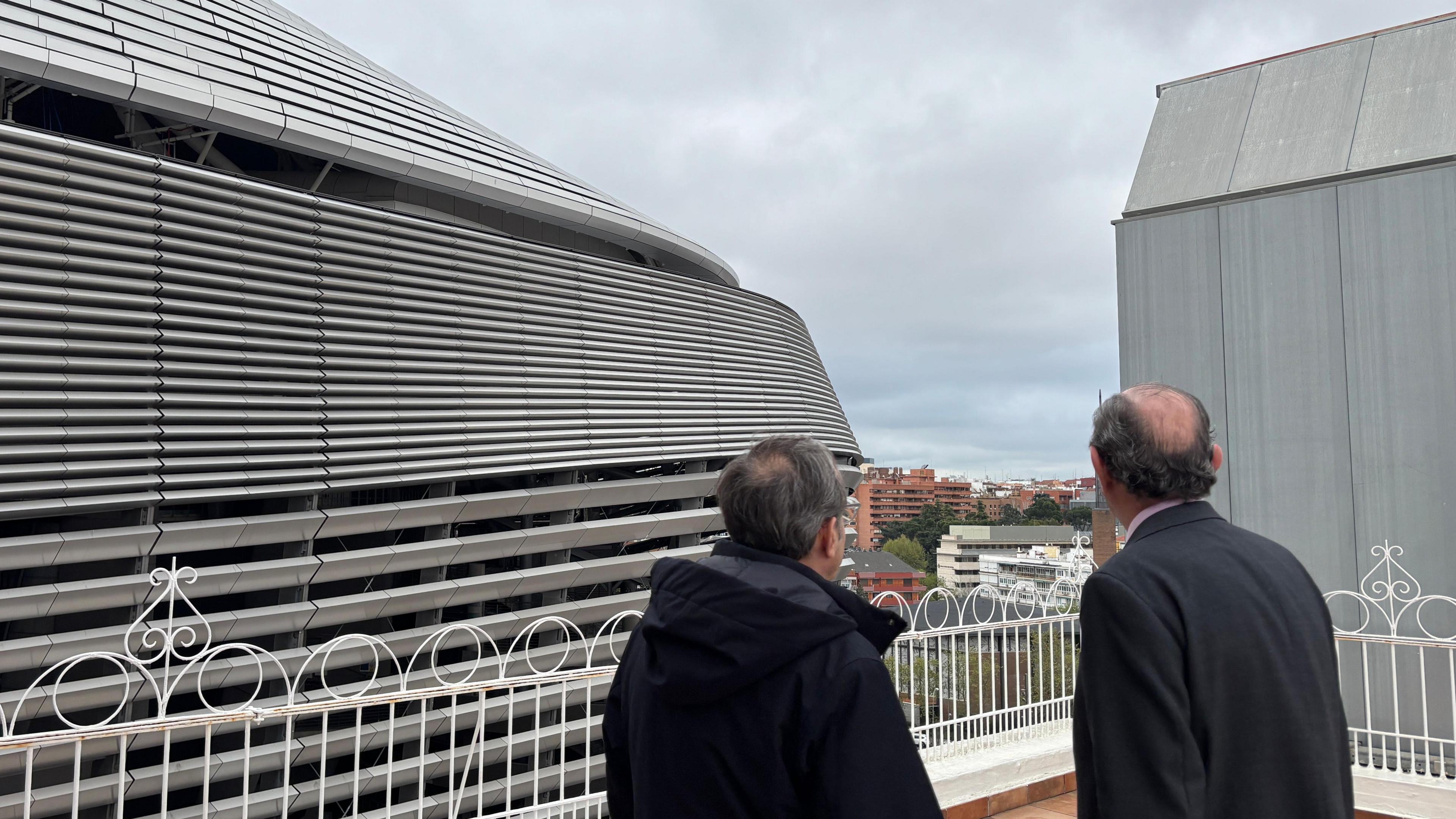 Two men stand with their back to the camera from the rooftop of a building looking over at the stadium which is right in front