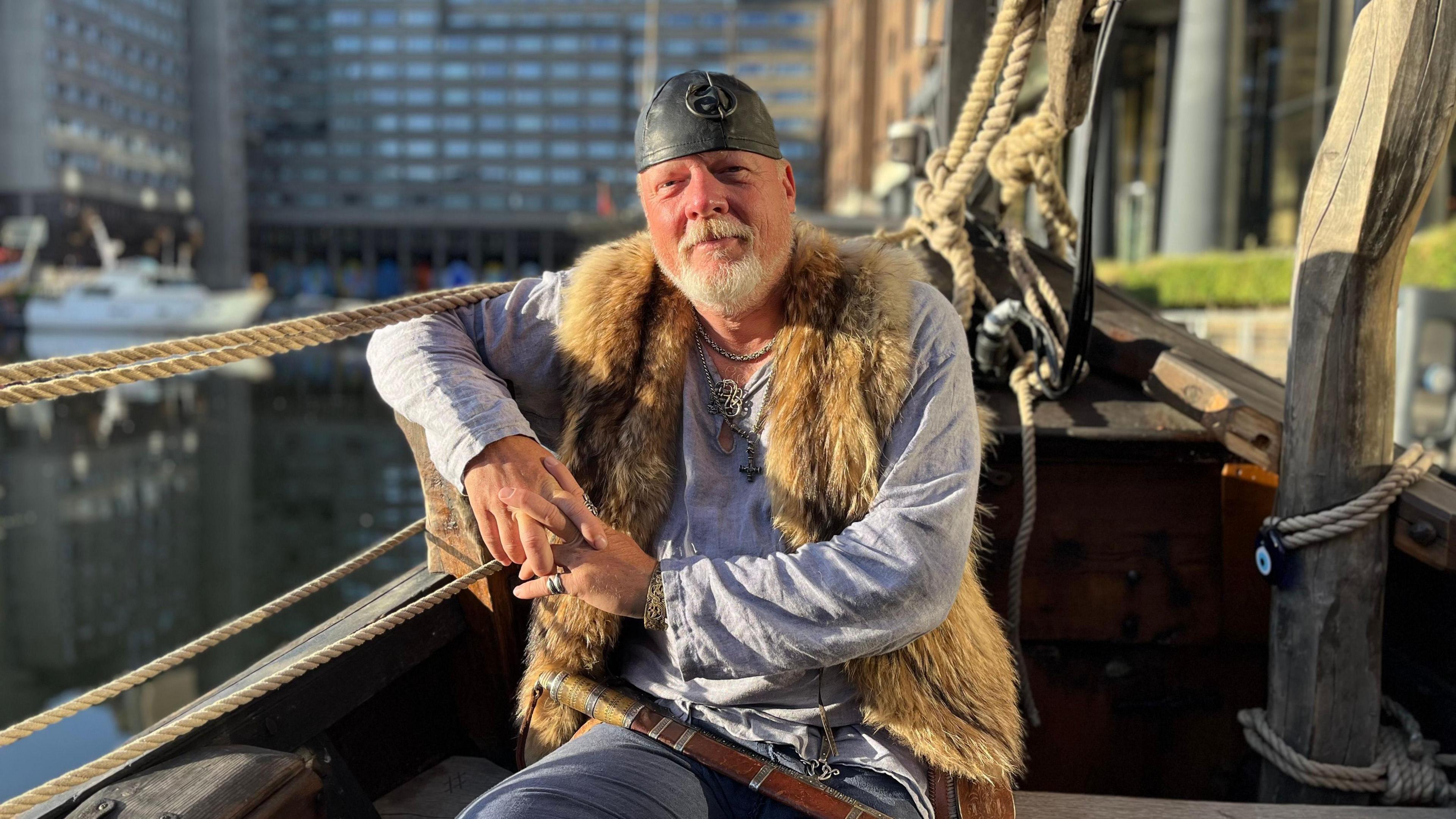 A man is sitting on a boat in dock, with his arm leaning on the rope along the edge of the boat. He is looking into the camera with a straight face, and has a white goatee beard. He is wearing a grey metal hat in the style of a Viking, and has a long, baggy grey top, with a fluffy brown vest on top.
