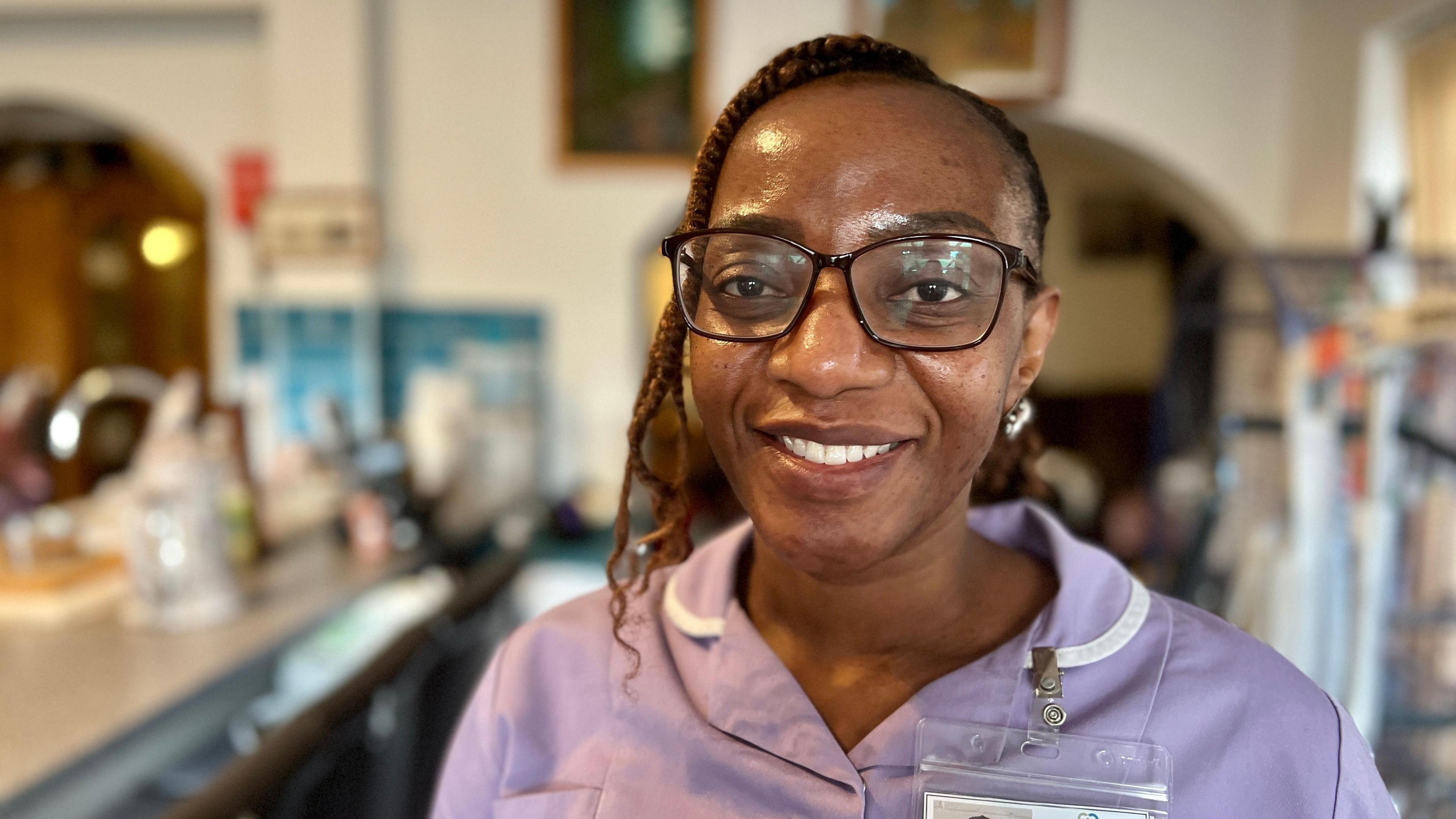 Care assistant Rhoda Machenga Mutimkulu looks at the camera, she is wearing large brown glasses and has her hair tied back. She is wearing a purple uniform with a name badge and is smiling, she is standing in someone's living room with a hoist in the background.