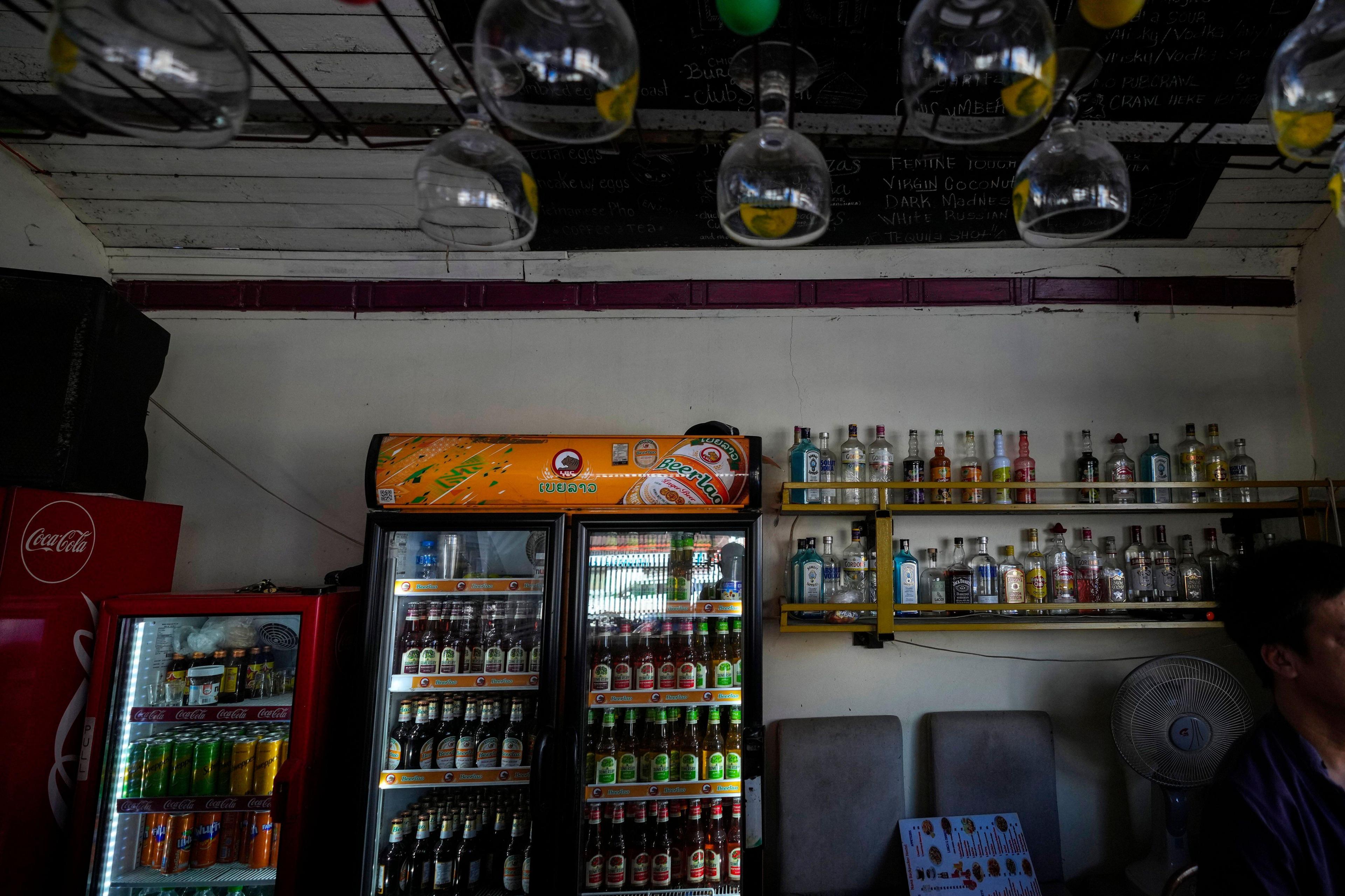 A man sits behind a bar which has a drinks fridge and shelves filled with bottles of beer and spirits