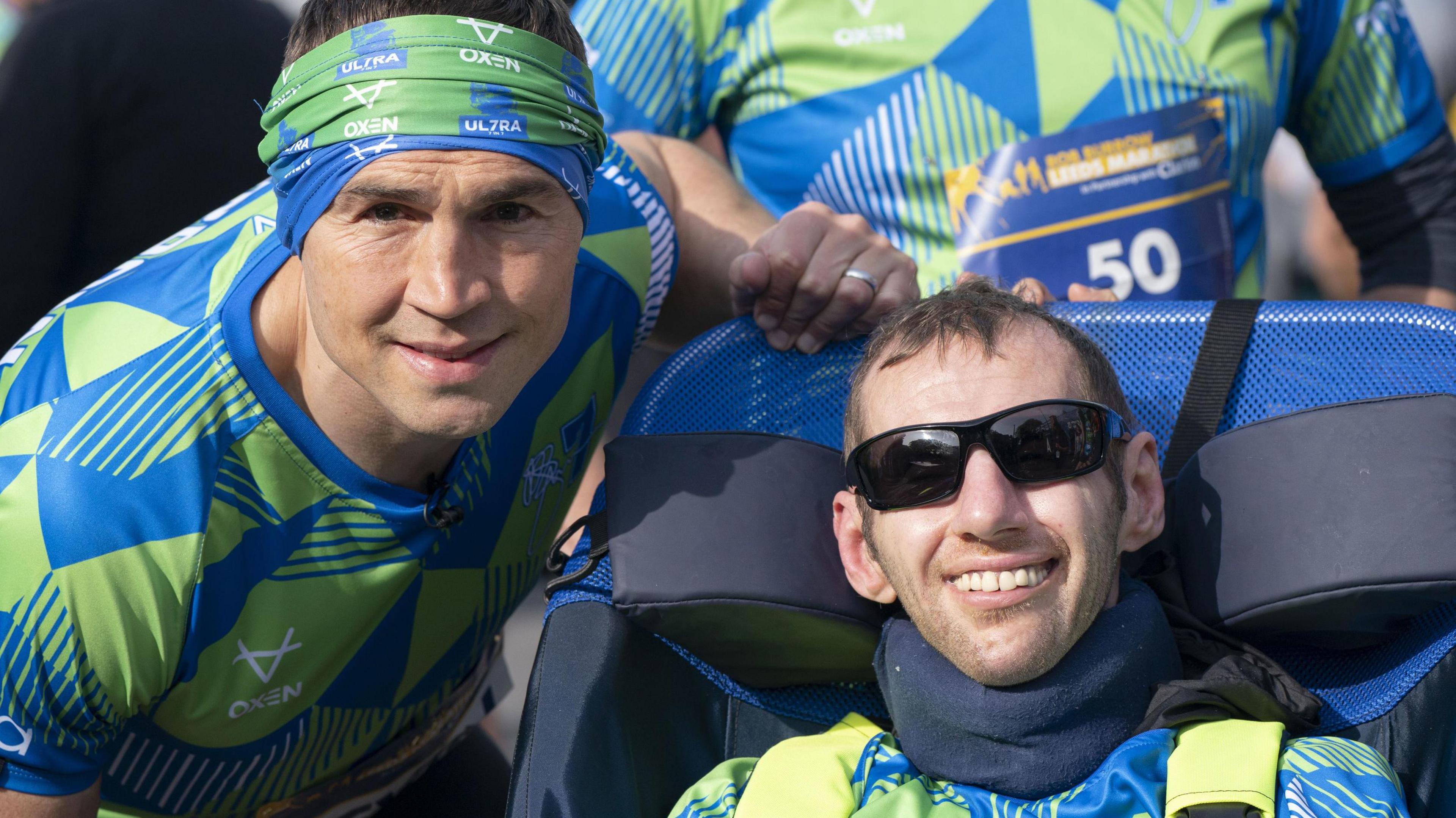 Kevin Sinfield wears a blue and green shirt and matching headband as he leans close to Rob Burrow, who is wearing the same top and sunglasses as he sits in a wheelchair. Both men are smiling and looking towards the camera.