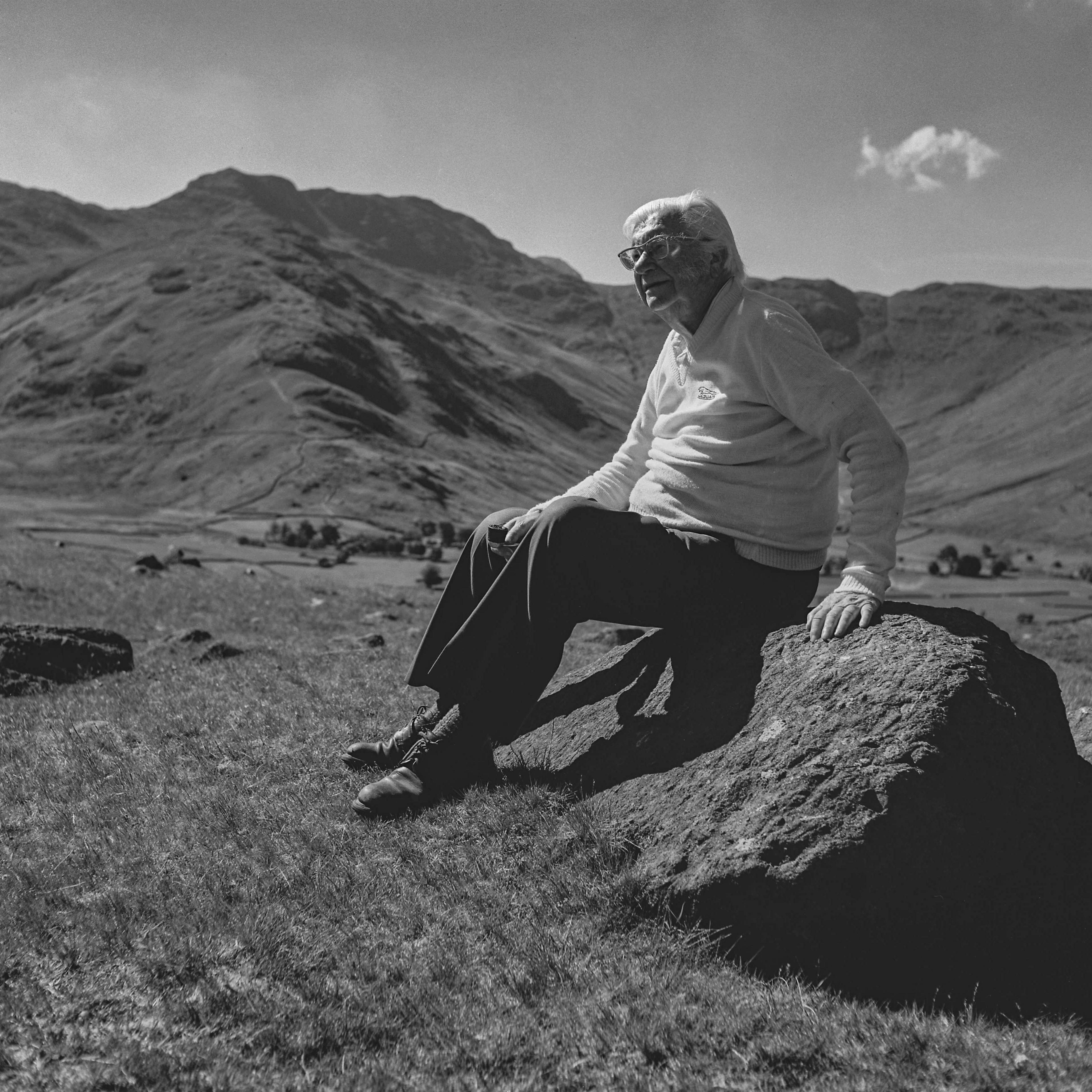 Black-and-white photograph of Alfred Wainwright sitting on a rock with the fells behind him. He is looking to the left of the image, and is wearing a light jumper and dark trousers. He has white hair and has glasses.