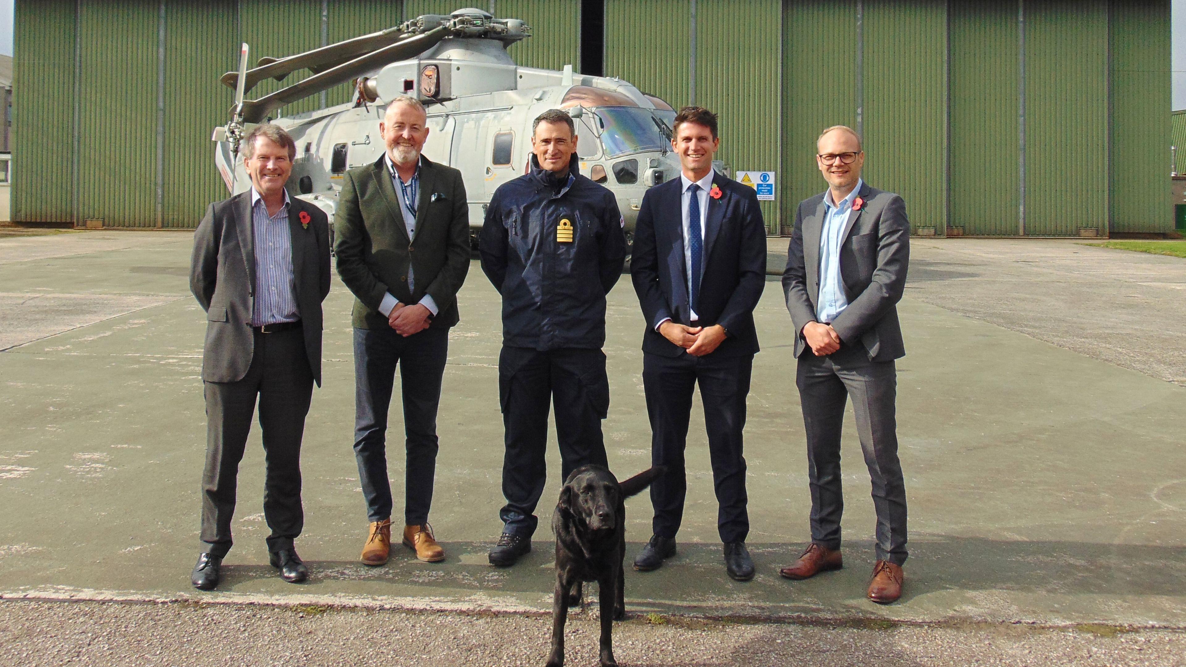 Outside a hangar at the military base. Five men are pictured. Four of those in the photo are wearing formal shirts with blazers. Captain Stuart Irwin is photographed in the centre wearing a military coat, navy blue in colour. They are photographed in front of a helicopter. 