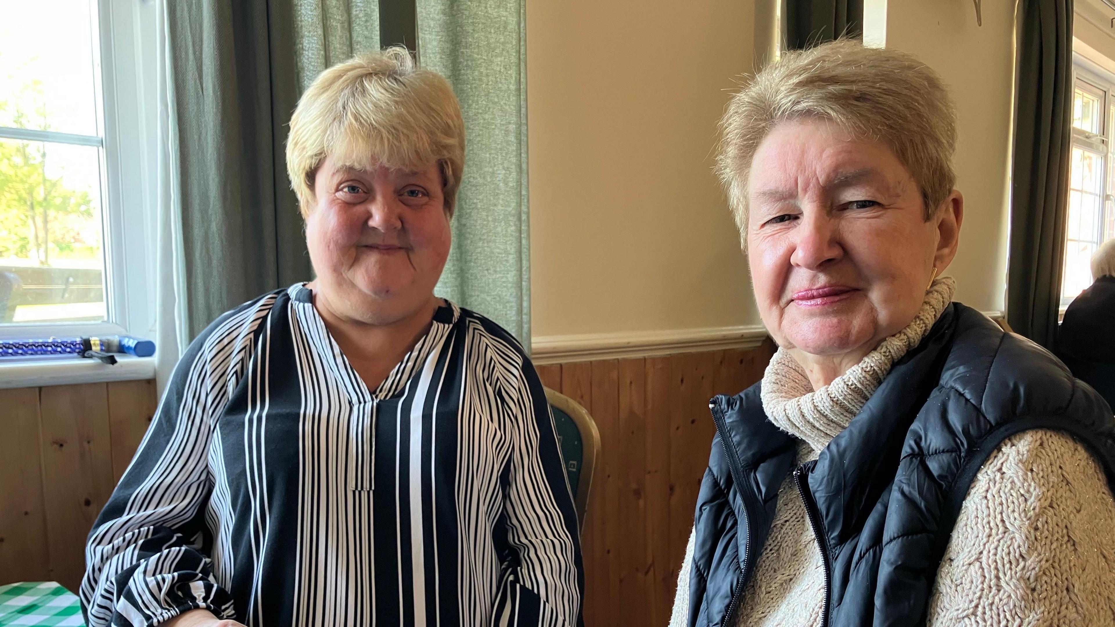 Joanna French and Dorothy Meeks smile at the camera while sitting at a table. Behind them is a window draped with green curtains. 